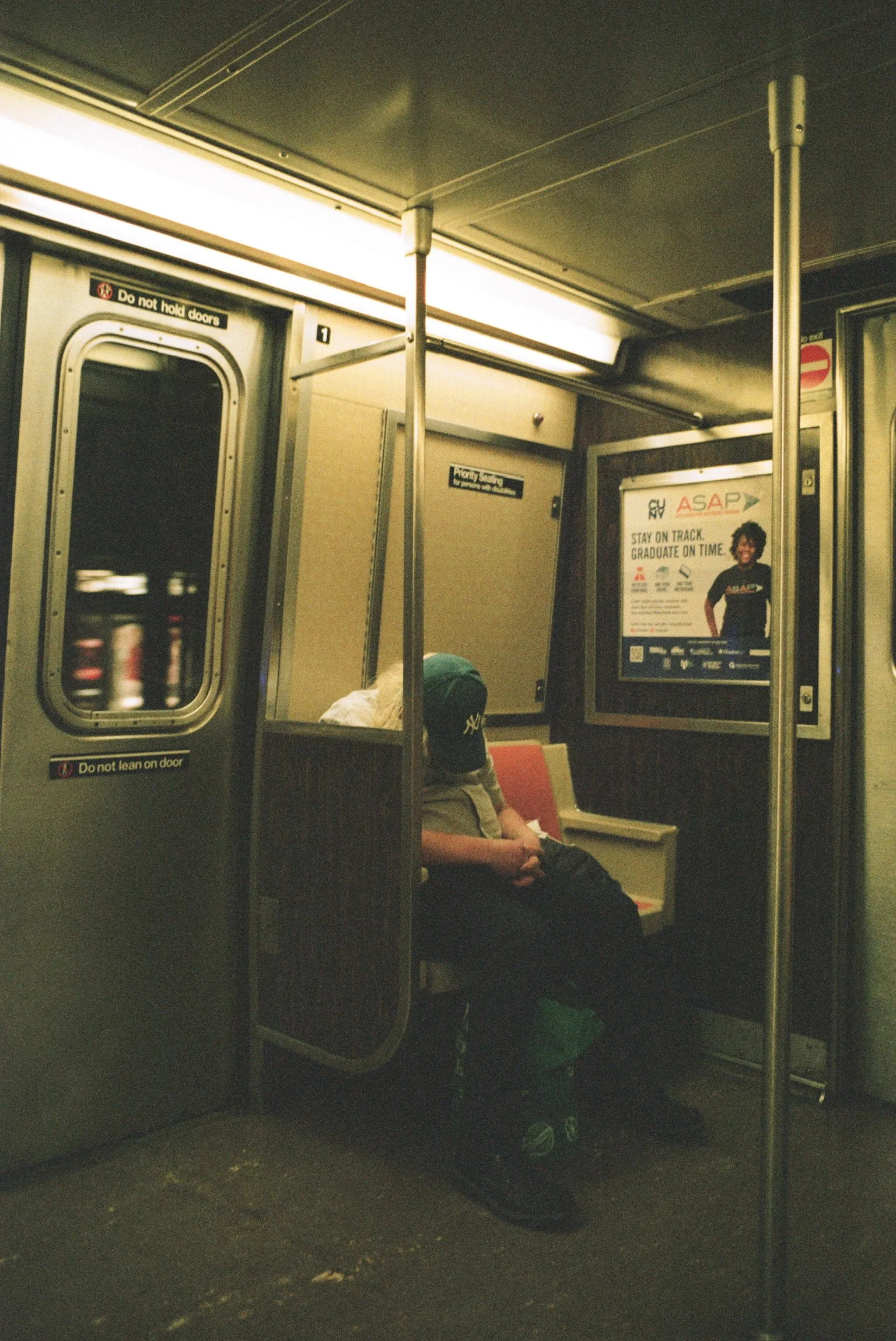 A person wearing a green baseball cap sitting on a train. The train has a yellowish seat, a pole, and signs on the walls, with a window showing blurred visuals outside.