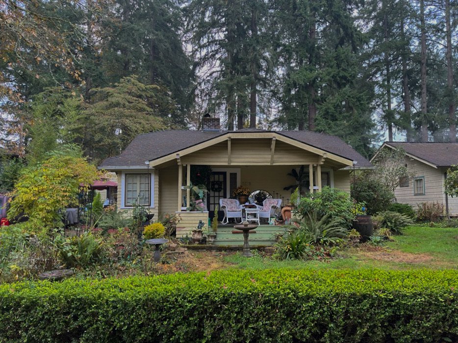 A cozy yellow house with a large front porch surrounded by a lush garden and tall trees.
