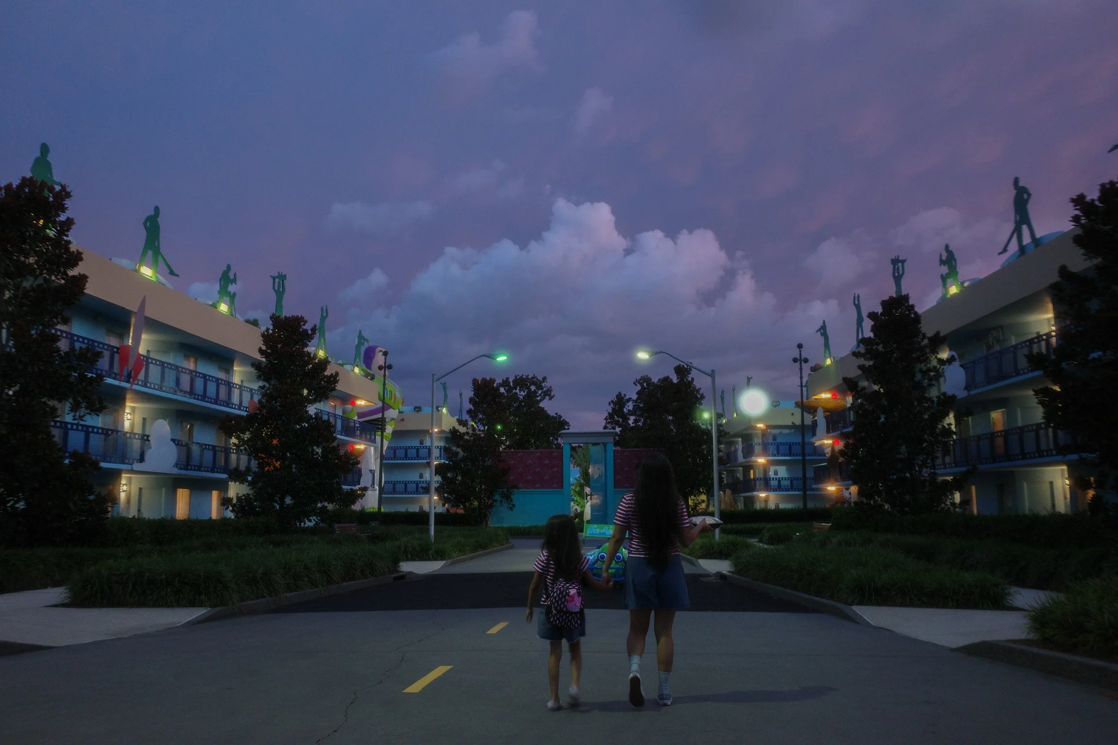 A woman and a young girl walking hand in hand on a paved road in front of a colorful, whimsical hotel decorated with various illuminated sculptures of people and other figures, during dusk with a dark blue sky and clouds.