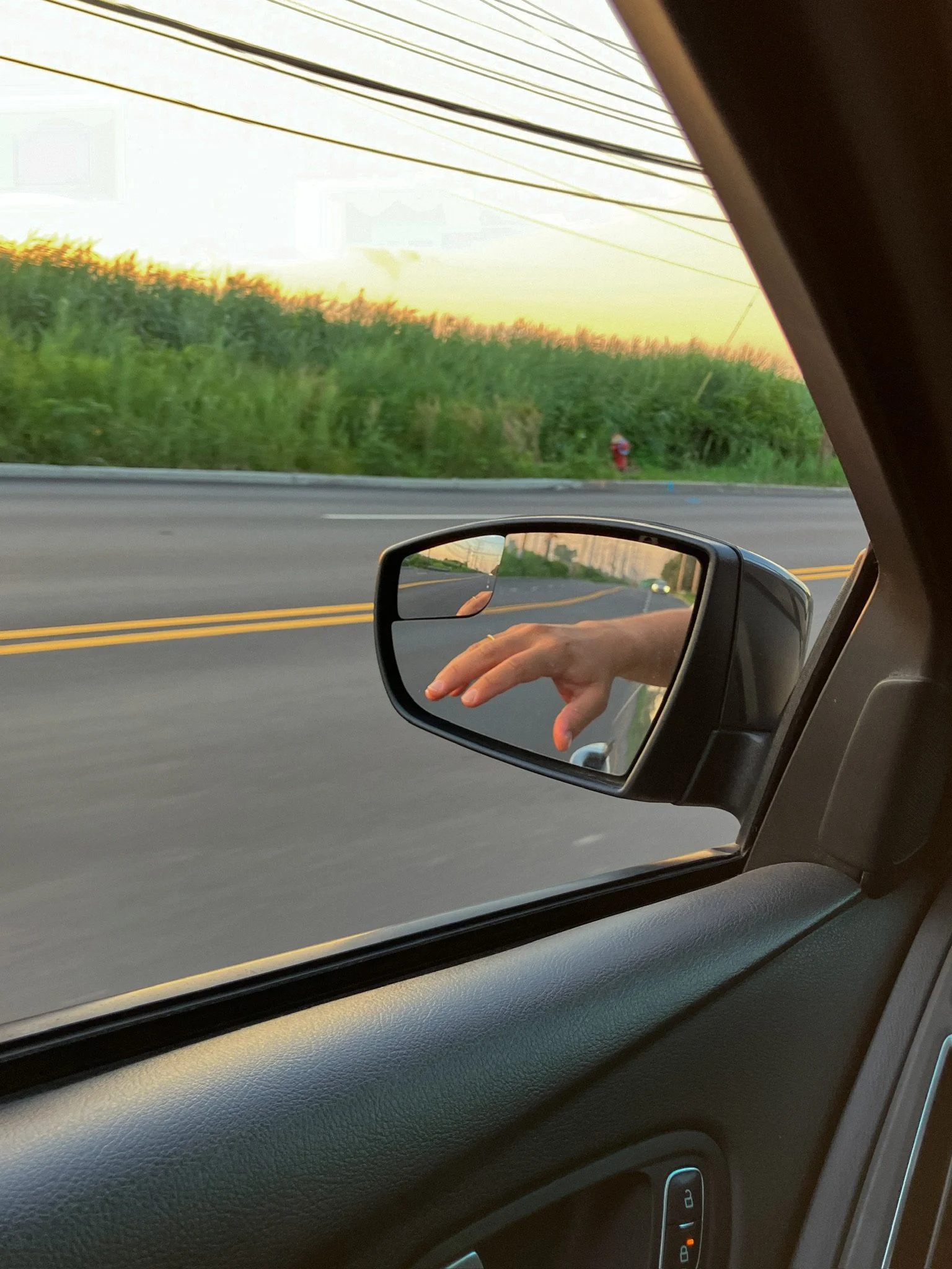 View from inside a moving car showing the side mirror reflecting a hand, empty road, and green vegetation with power lines and sunset sky in the background.