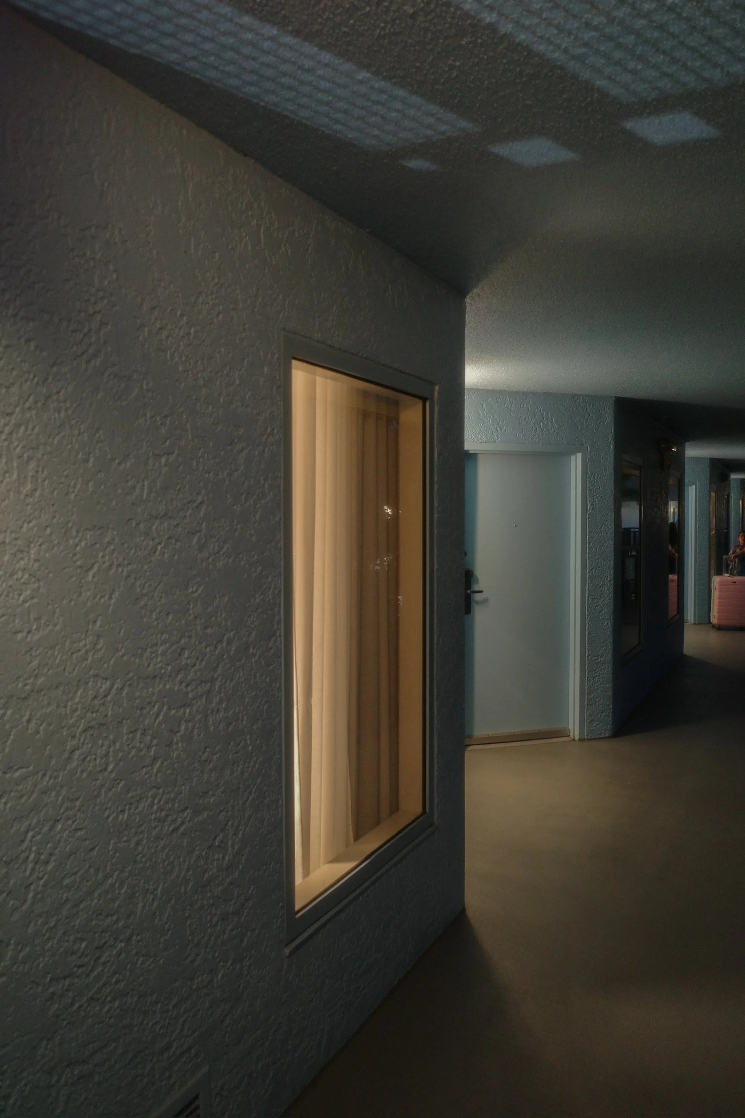 Hotel corridor with textured gray walls, closed white door, and window with blinds showing interior curtains. Dim lighting.
