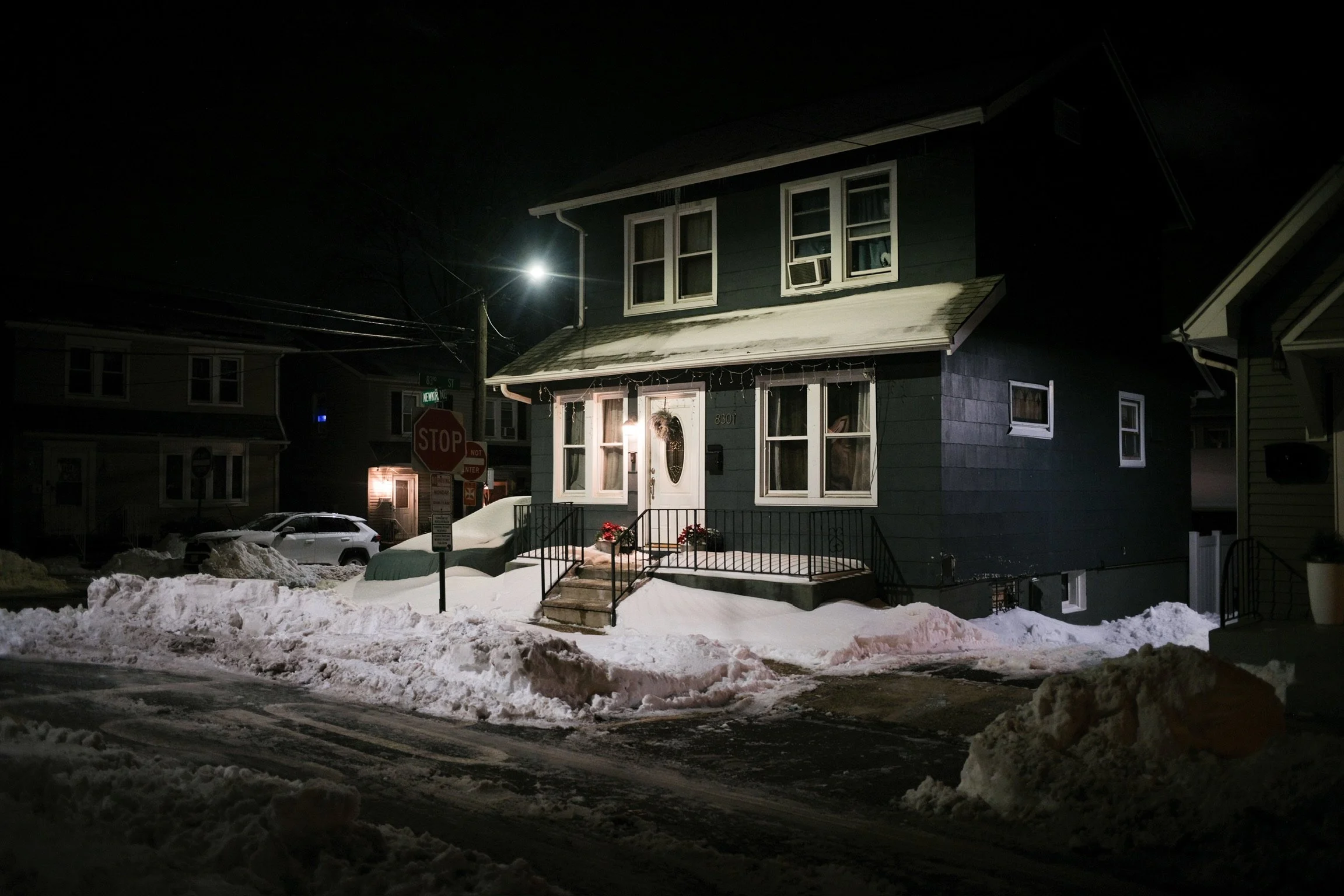 A dark nighttime scene of a blue two-story house with a snow-covered roof and steps, illuminated by a porch light. There are snow piles along the street and parked cars nearby.