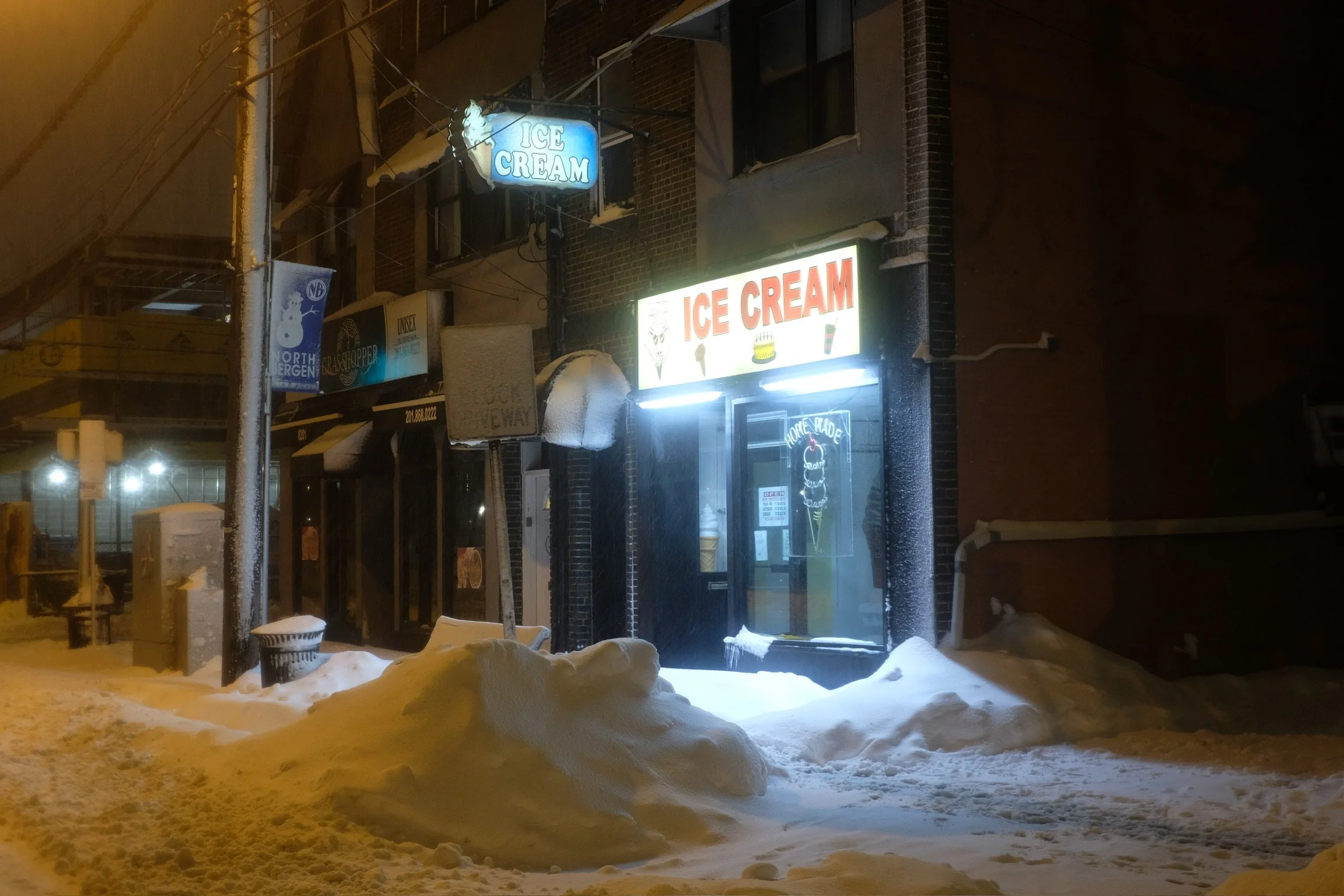 Snow-covered sidewalk outside a small ice cream shop at night with illuminated 'Ice Cream' signs, snow piled near the entrance, and snow falling.