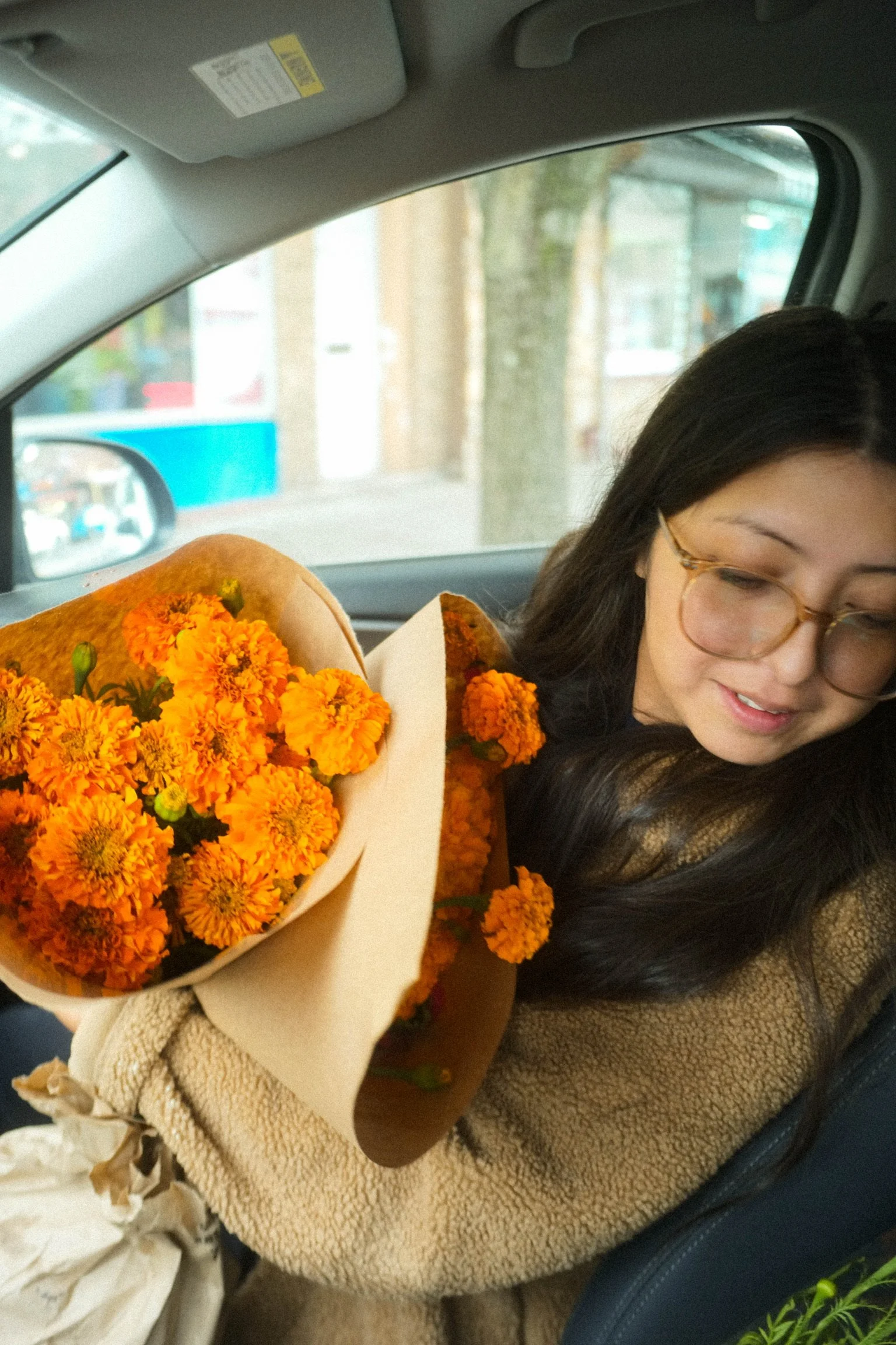 A woman with glasses holding a bouquet of orange marigold flowers inside a car.