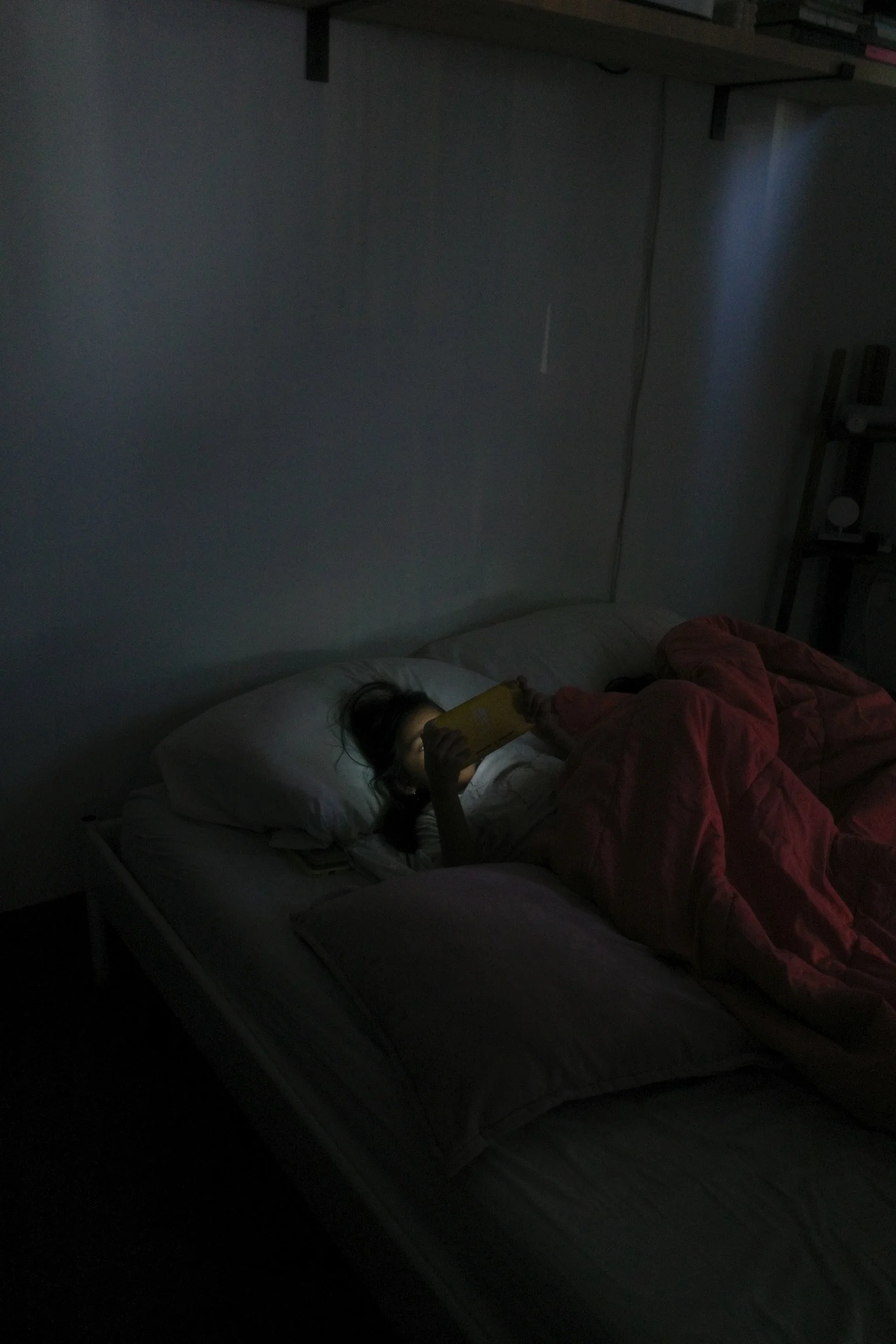 A person lying in bed at night, illuminated by the light from their phone screen, with a blanket over them, in a dark room.