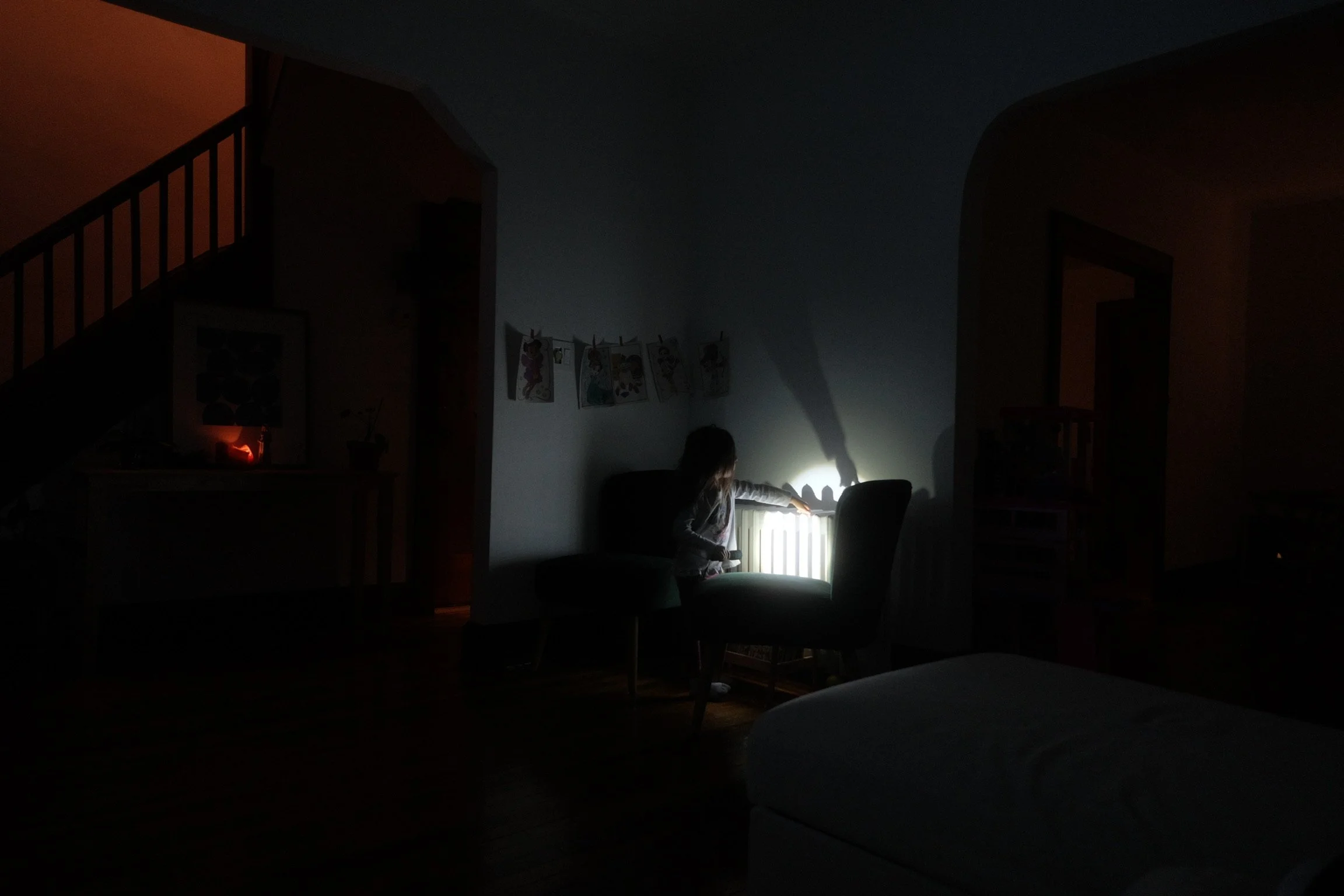 A young girl in pajamas reaching toward a radiator with a flashlight in a dark living room, illuminated by a small light on a side table and a string of artwork on the wall.