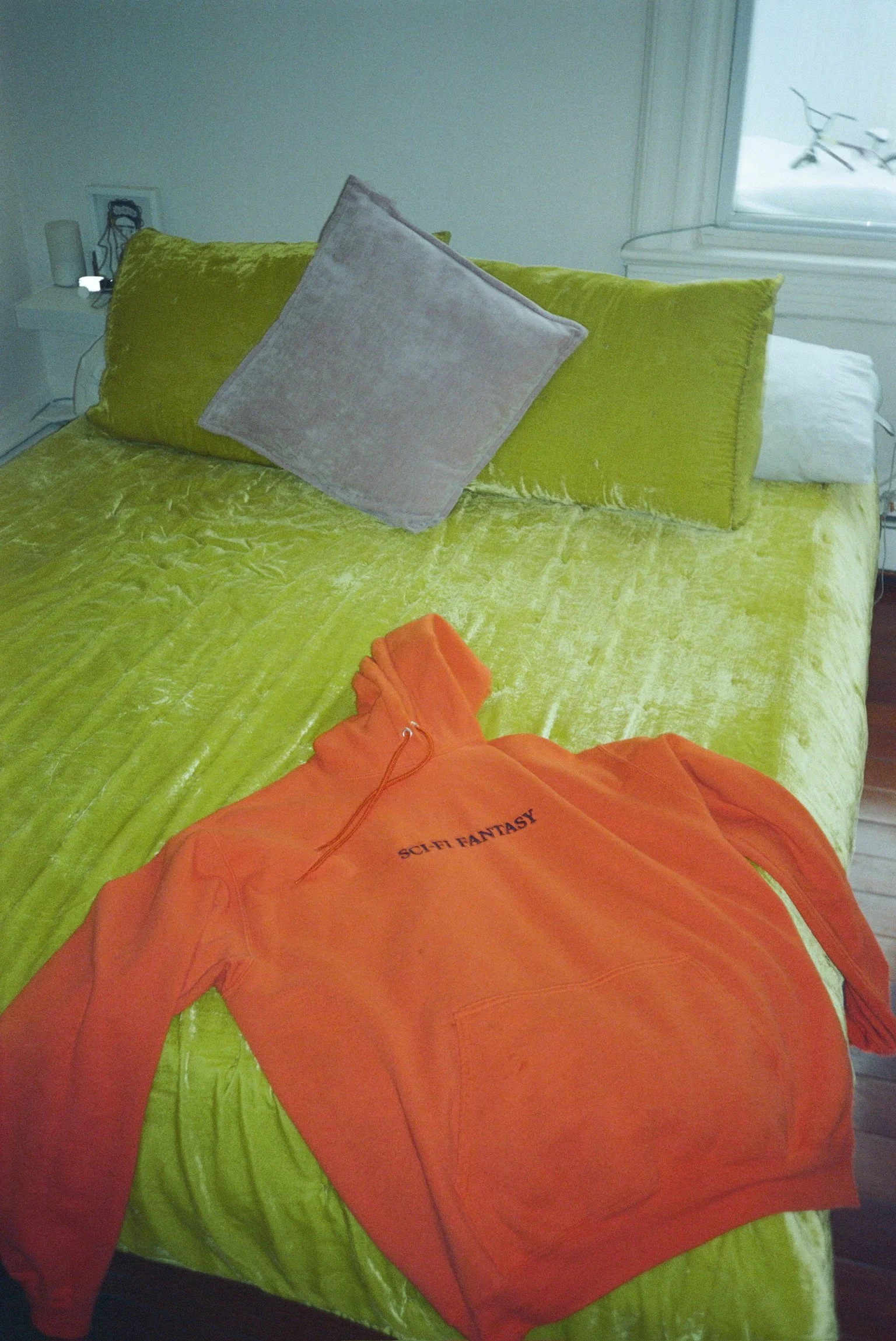 A bed with yellow-green velvet bedding, a white pillow, and a pink pillow, with an orange hoodie with 'SCI-FI FANTASY' printed on it laying on the bed, and a nightstand with a lamp and picture frame beside it.