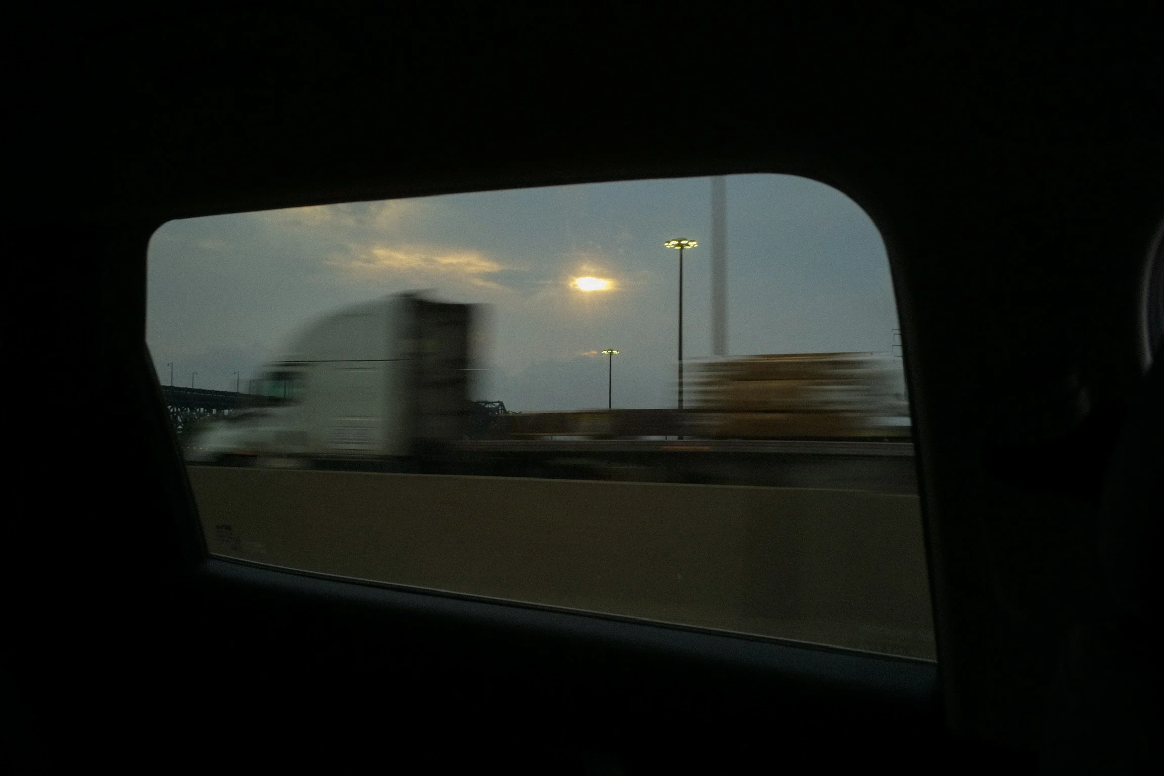 View of a highway through a car window, with a blurred truck passing by and the sun partially obscured by clouds during dusk or dawn.