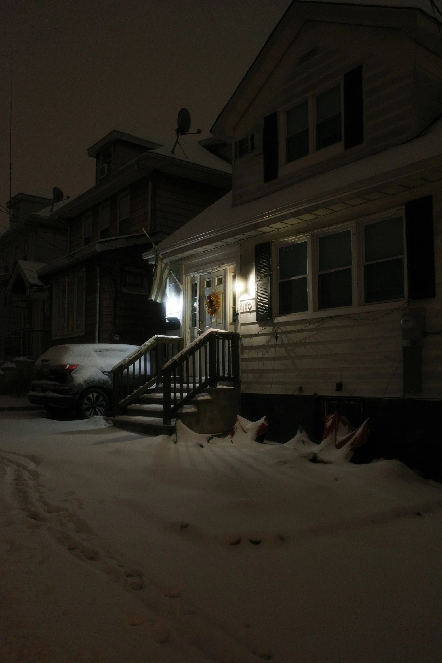 A snowy night scene of a residential house with exterior lights on, decorated with a holiday wreath on the front door. A car is parked in front of the house, covered in snow, along with the snow-covered ground and footprints visible in the snow.