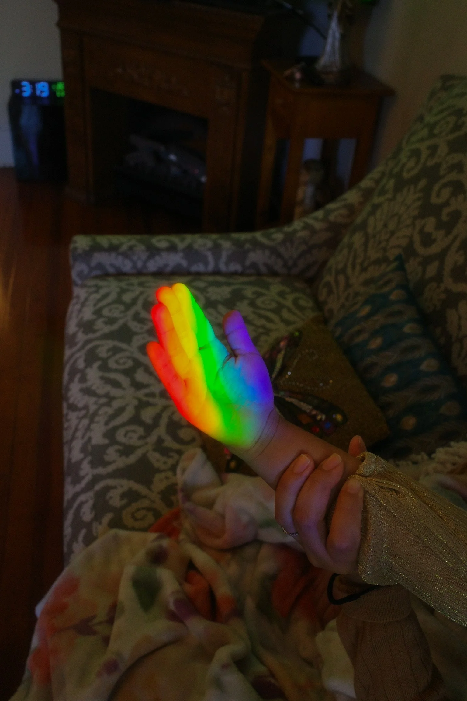 A person's hand is raised with a rainbow-colored light projected on it, showing a spectrum of colors from red to violet. The background includes a patterned chair, a wooden table, and a small dog partially visible under the table.