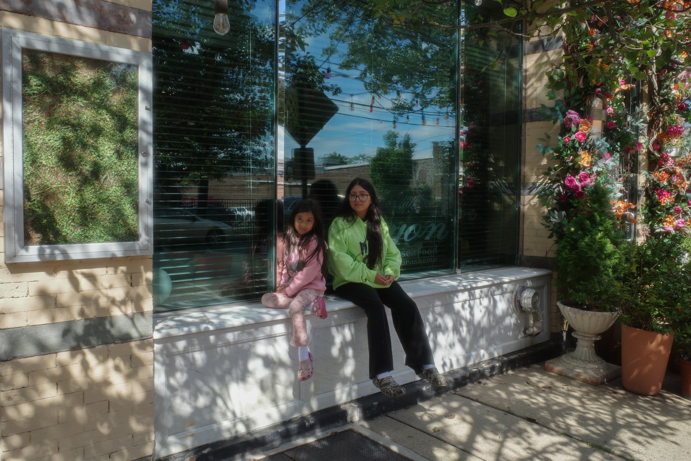 Two girls sitting on a white ledge outside a glass storefront, with trees casting shadows on the sidewalk and potted plants nearby.