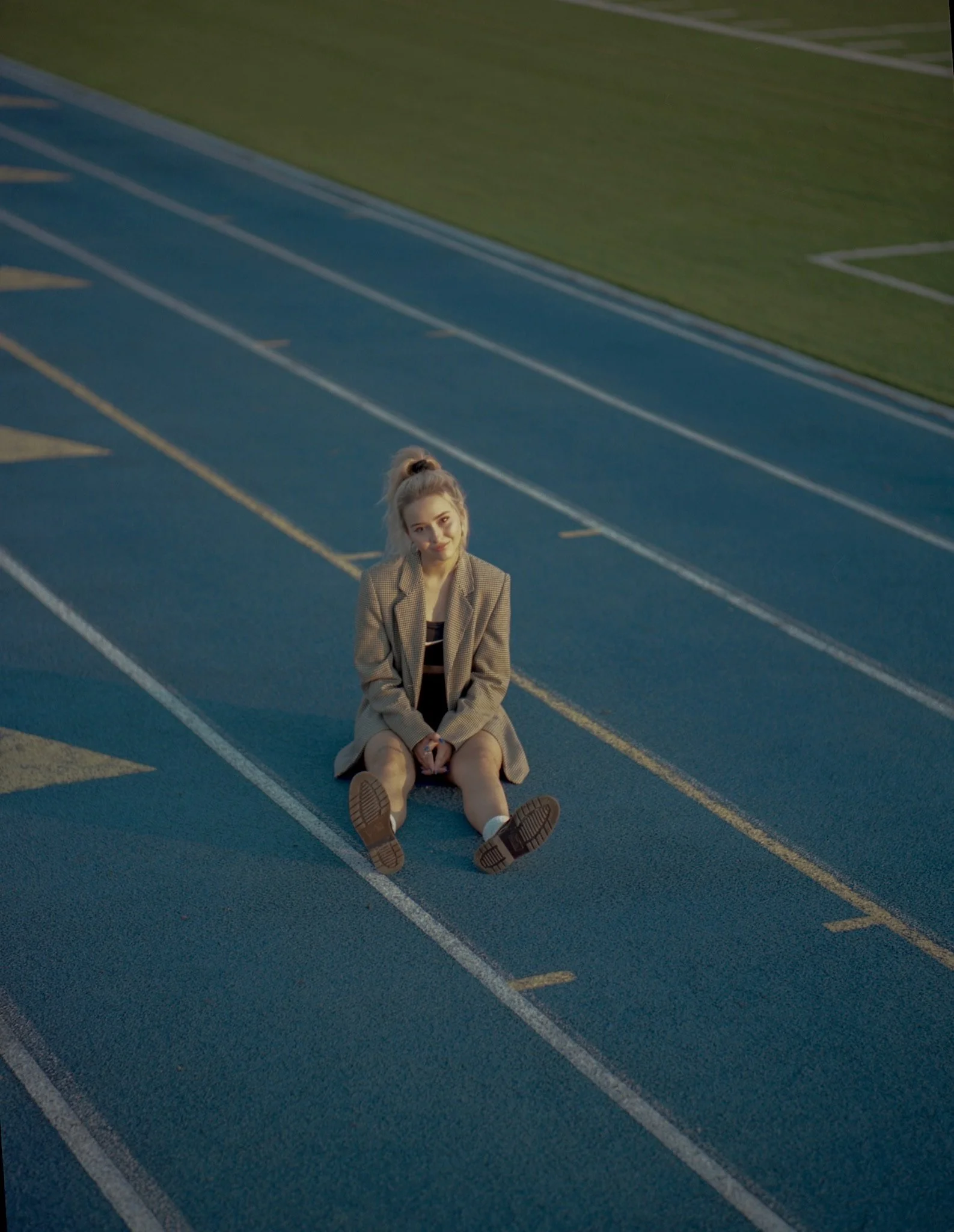 A young woman sitting on a blue running track with painted lines, wearing a brown checkered blazer and black dress, smiling at the camera.