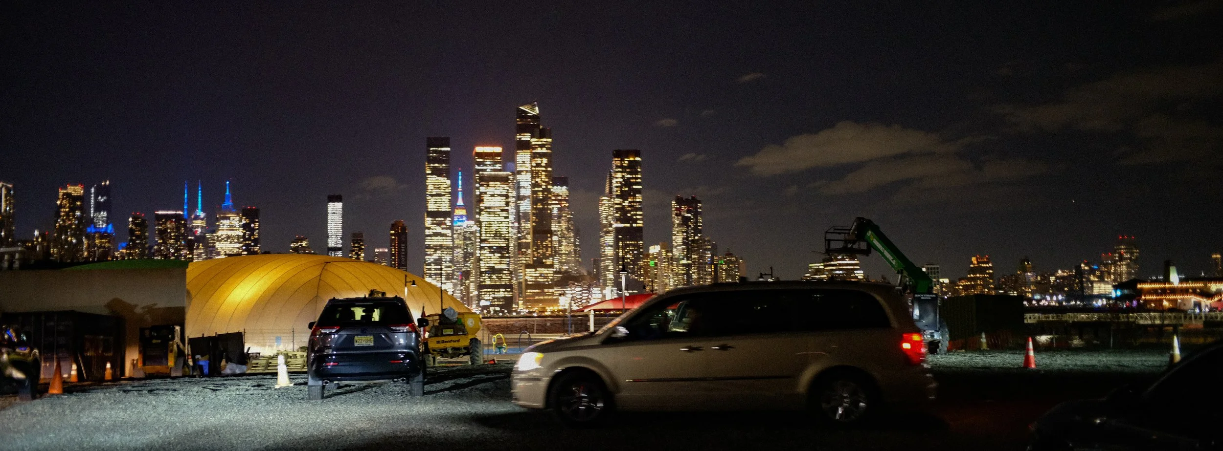 Nighttime cityscape of a downtown skyline with illuminated skyscrapers, seen from a parking lot with parked cars and construction equipment in the foreground.