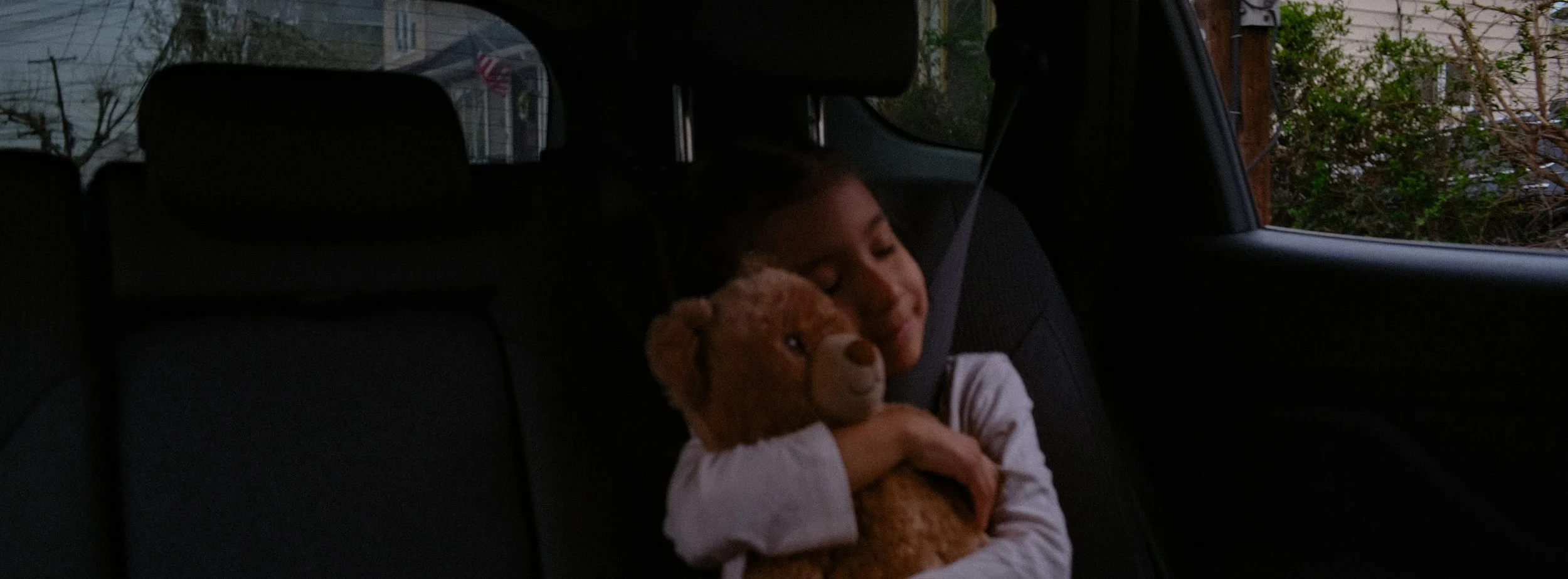 A young girl sleeping in the backseat of a car, hugging a stuffed animal.