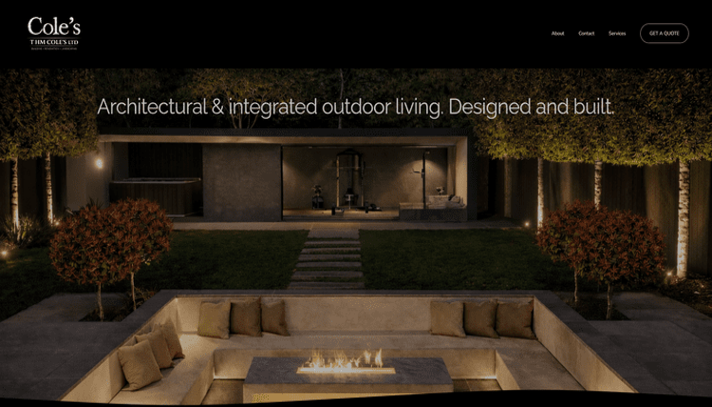 Night view of a modern outdoor living space with a built-in fireplace, surrounded by a seating area with cushions, trees, and landscape lighting.