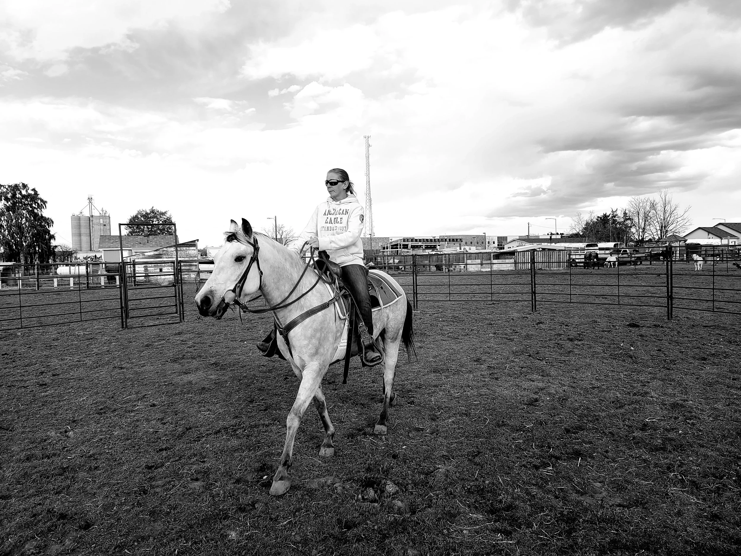 A woman riding a horse in an open outdoor riding arena with fences, under a cloudy sky.