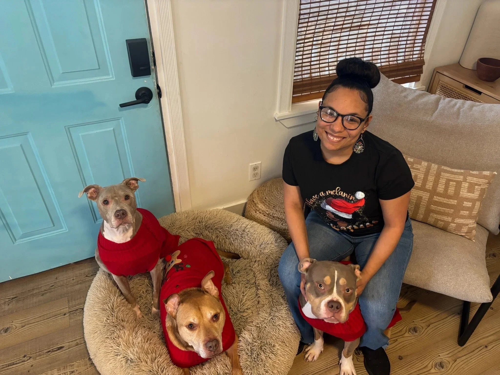 A smiling woman wearing glasses and a black Christmas sweater with a Santa hat design, sitting on a chair with three dogs dressed in red sweaters, one on a dog bed and two on the floor, in a cozy indoor setting with a blue door and wooden floor.