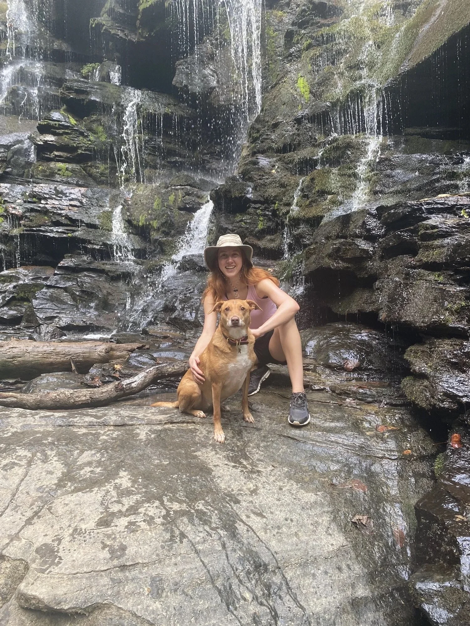A woman and a dog posing in front of a small waterfall in a forested area, with rocks and moss around them.