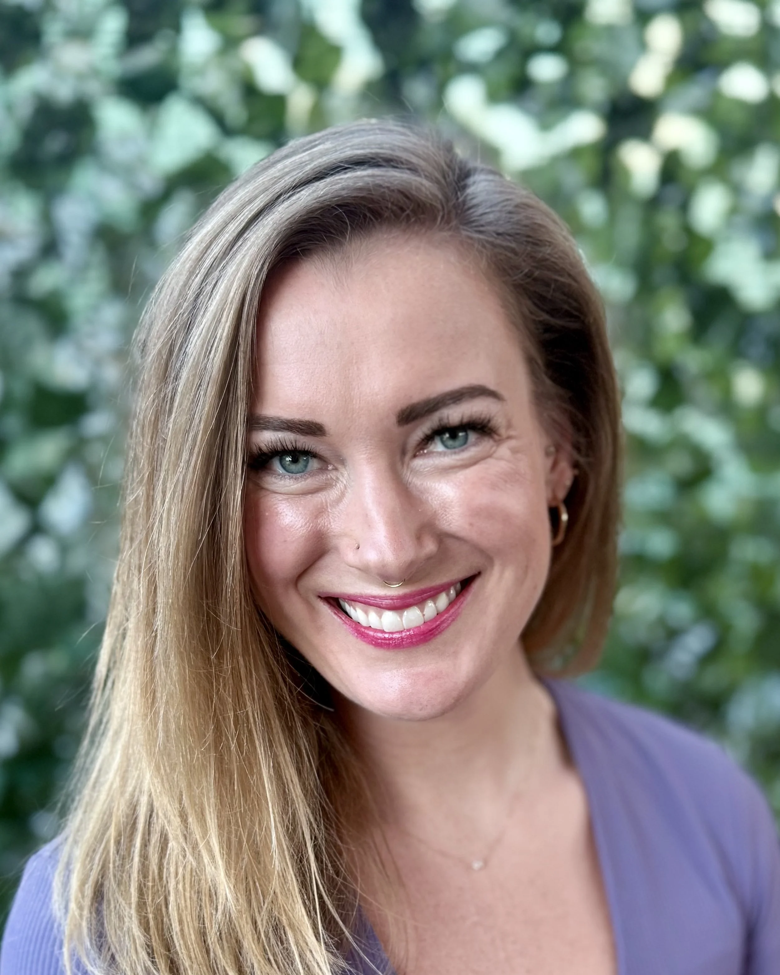 A smiling woman with long blonde hair and blue eyes, wearing a purple top and a small necklace, outdoors with green leaves in the background.