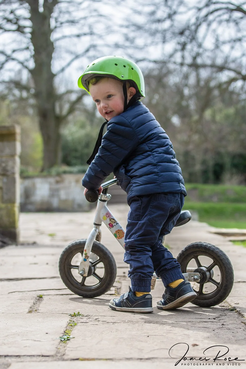 A young boy with a green helmet riding a small balance bike on a paved outdoor path, wearing a blue puffy jacket and dark pants.