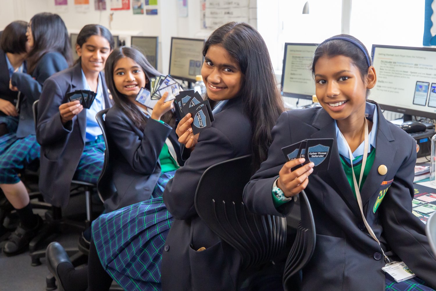 Group of young girls in school uniforms sitting at desks with computers, holding cards related to cyber security.