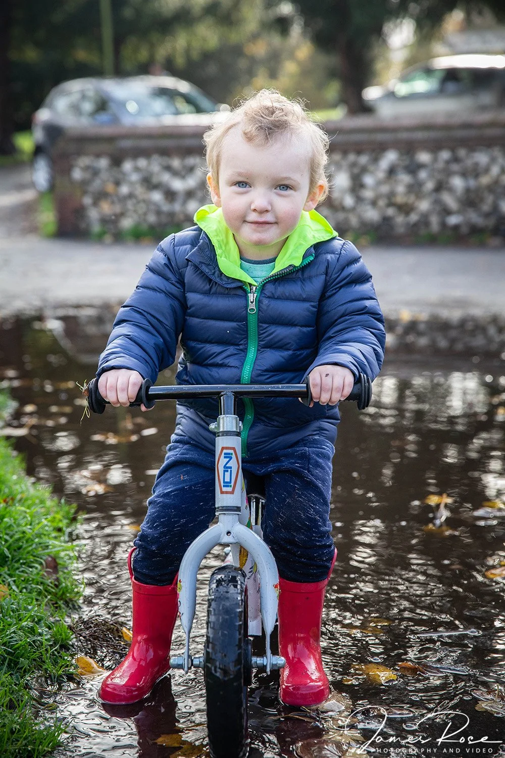 A young child wearing a blue jacket, red rain boots, and a bright green hoodie rides a small bike through a shallow puddle in a park or outdoor area.