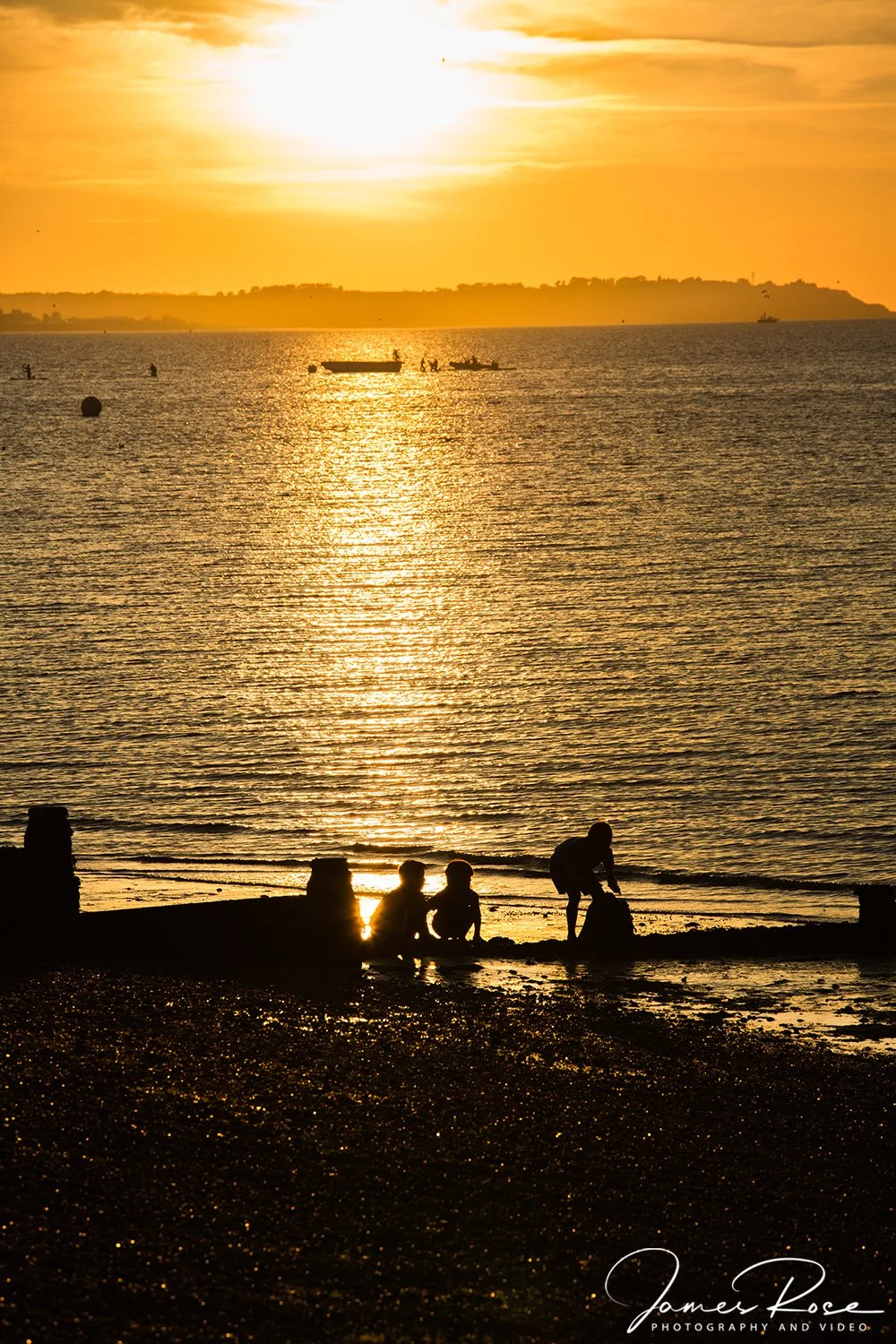 Silhouettes of children playing on a beach at sunset, with boats on the water and a cloudy sky illuminated by the setting sun in the background.