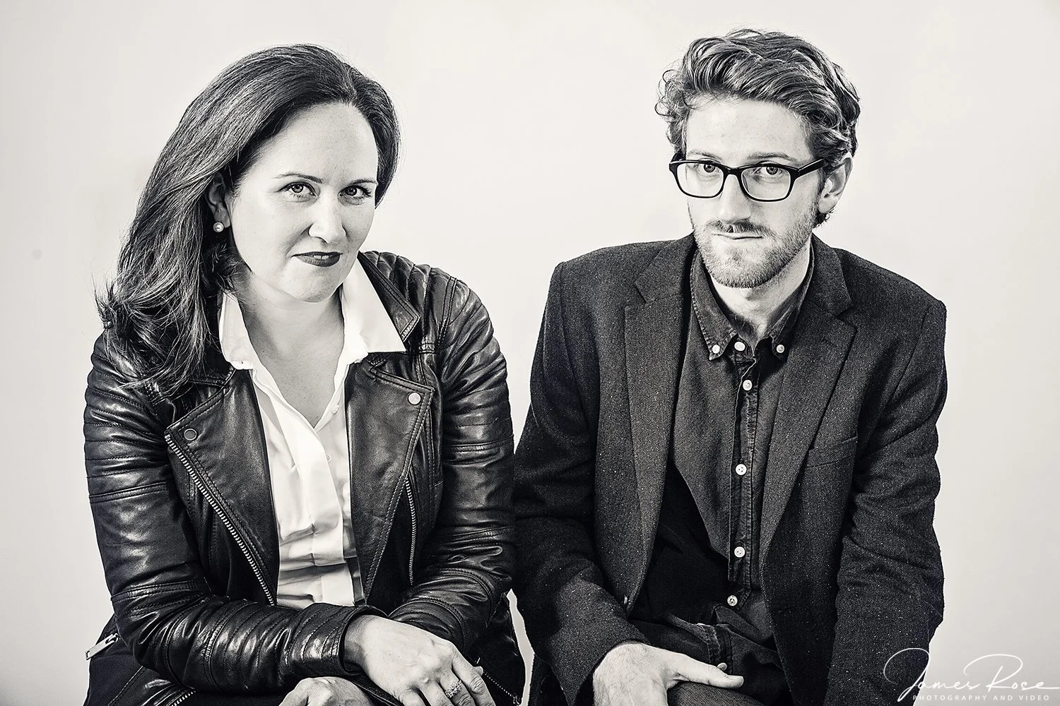 A black-and-white photo of a woman and a man sitting together, both dressed in dark clothing; the woman is wearing a leather jacket and a white shirt, and the man is wearing glasses and a blazer.