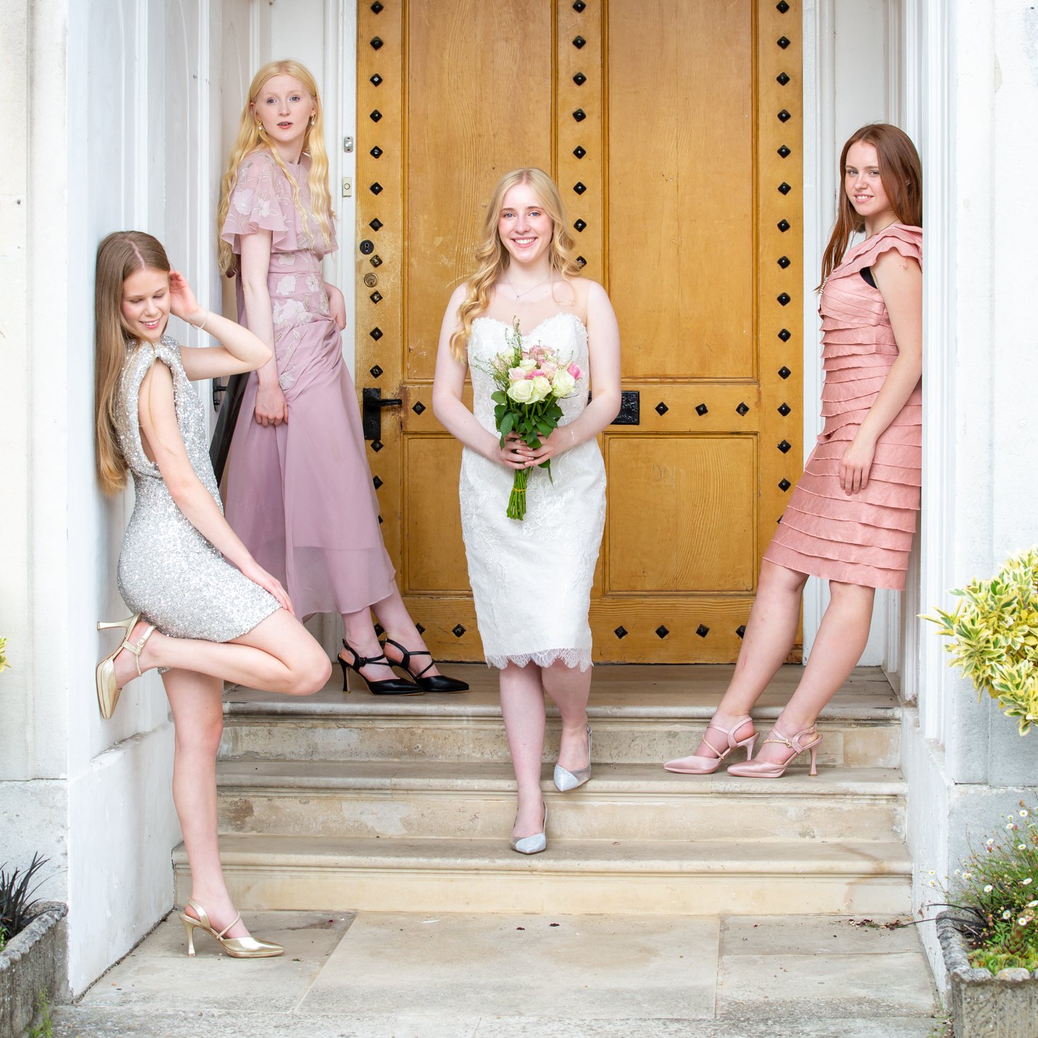A bride in a white lace dress holding a bouquet of white and pink flowers, standing on front stairs with four bridesmaids dressed in pink and silver, all smiling and posing in front of a wooden door.