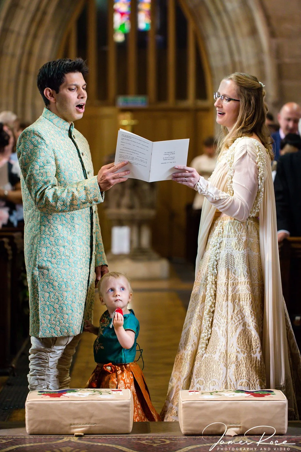 A couple exchanging vows or rings during a wedding ceremony inside a church with stained glass windows. The man is dressed in an ornate cream and gold jacket, and the woman in a long, decorative dress. A child stands in front of them, looking up.]