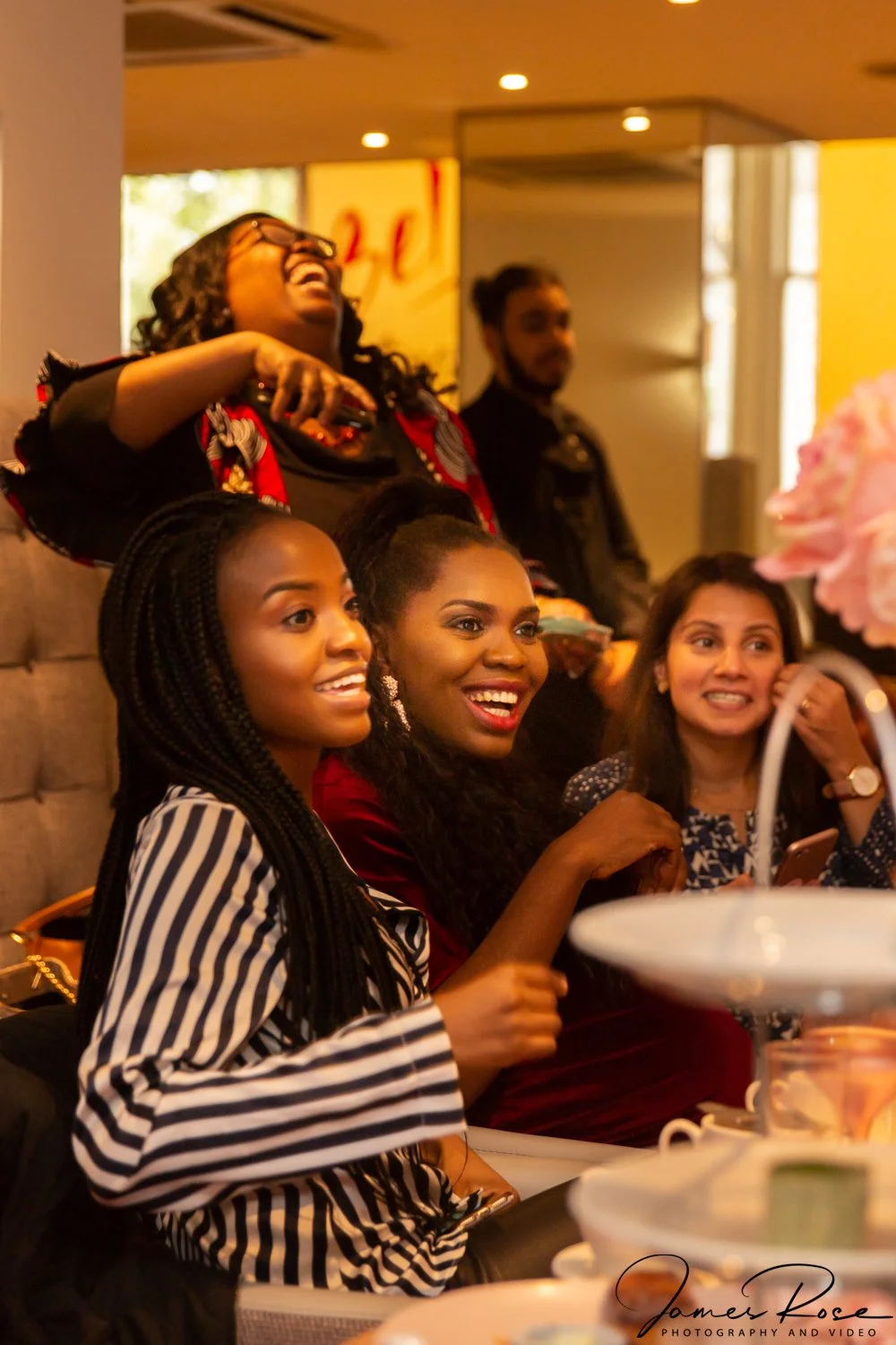 A group of diverse women laughing and smiling at a social gathering at a table with desserts in a warmly lit restaurant or cafe.