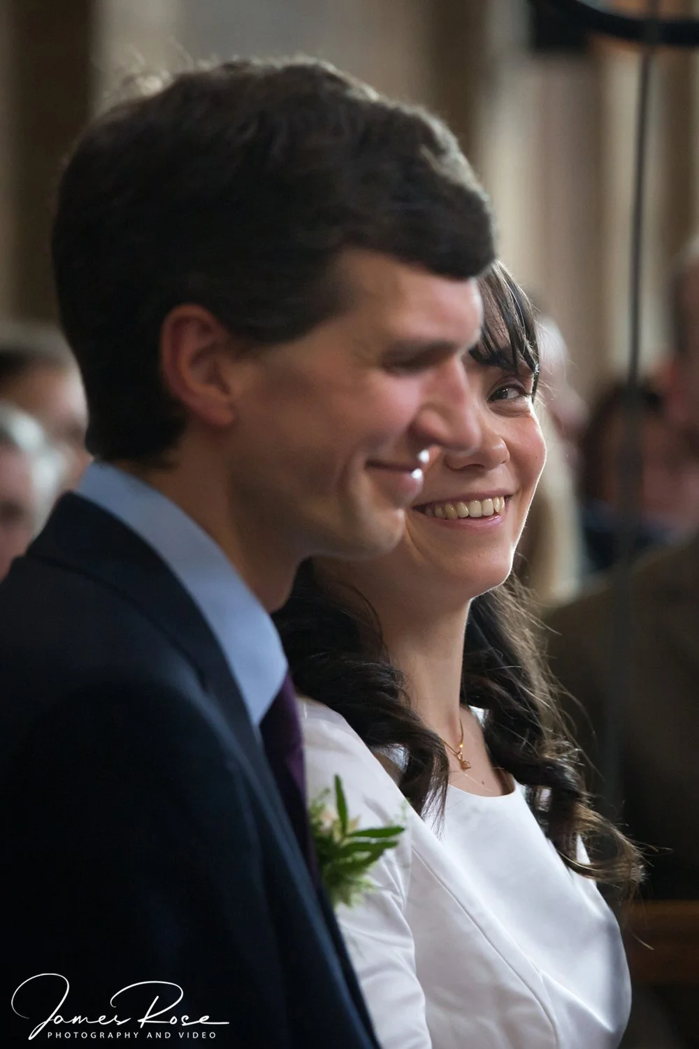 A man and woman are seated side by side at a formal event, smiling and looking toward the front. The man has dark hair and is wearing a dark suit with a light shirt and a tie, while the woman has dark hair and is wearing a white dress.