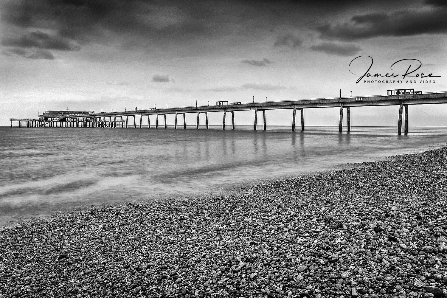 Black and white photograph of a pebble beach with a long pier extending into the ocean under a cloudy sky, with calm water and distant clouds.