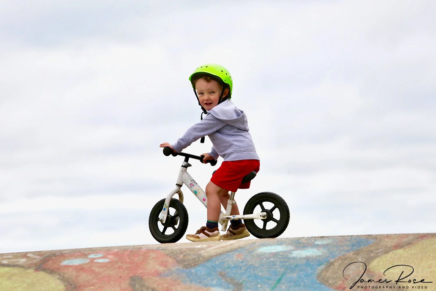 A young boy wearing a green helmet, gray hoodie, red shorts, and brown shoes riding a small white balance bike on a colorful concrete surface under a cloudy sky.