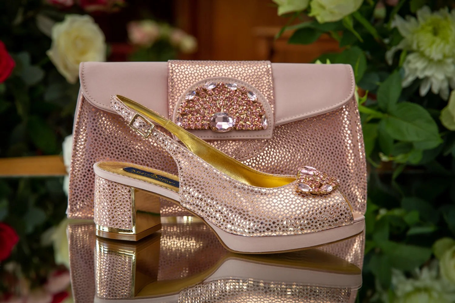 A pair of pink, glittery, open-toe high-heeled shoes with jeweled embellishments, placed in front of a matching pink clutch with similar jeweled design, on a reflective surface with a background of green and white flowers.