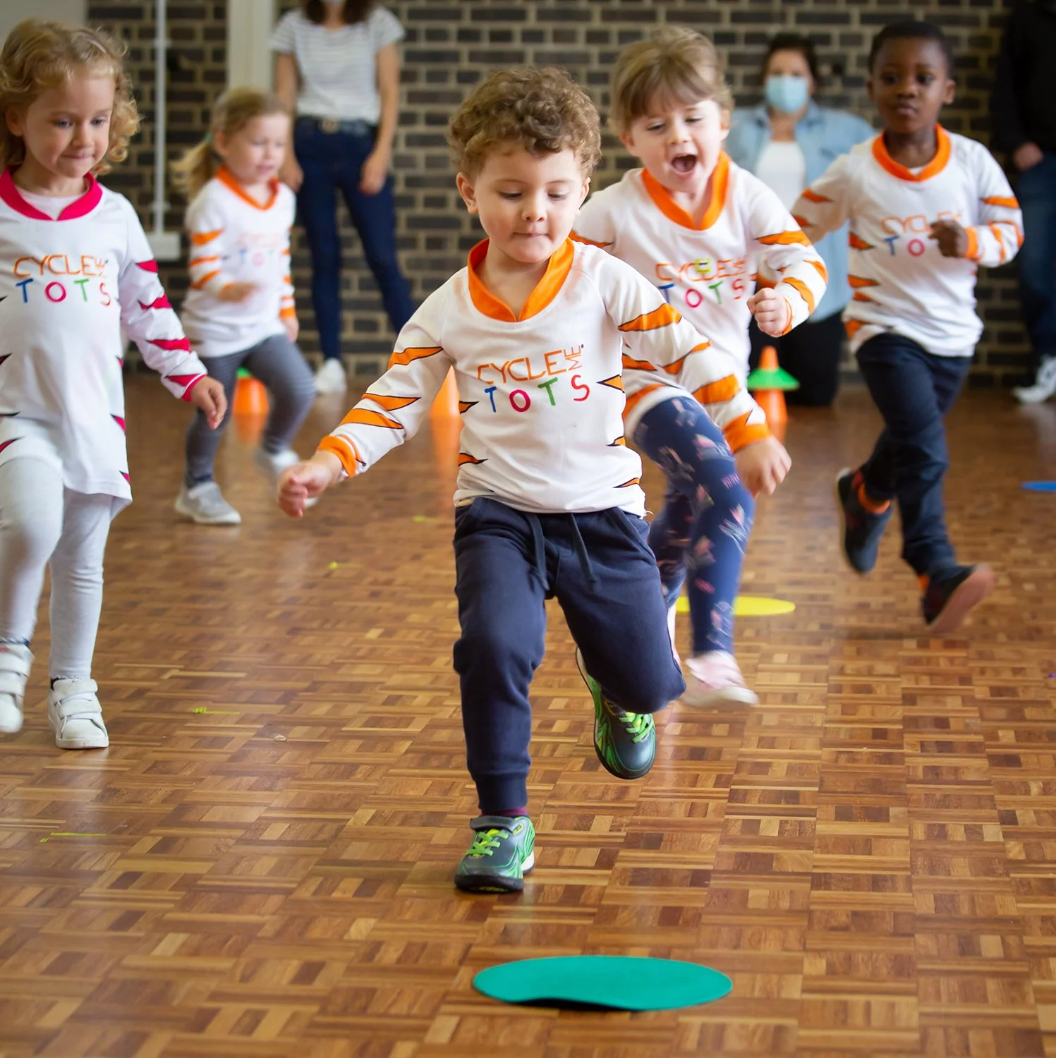 Children participating in an indoor activity, jumping over small colored cones on a wooden floor, wearing matching 'Cycle Kids' shirts.
