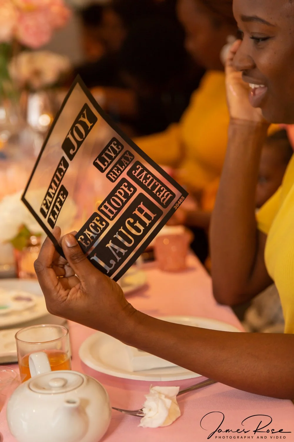 Woman in yellow dress reading a sign with positive words at a table with a teapot, glass, and plates.