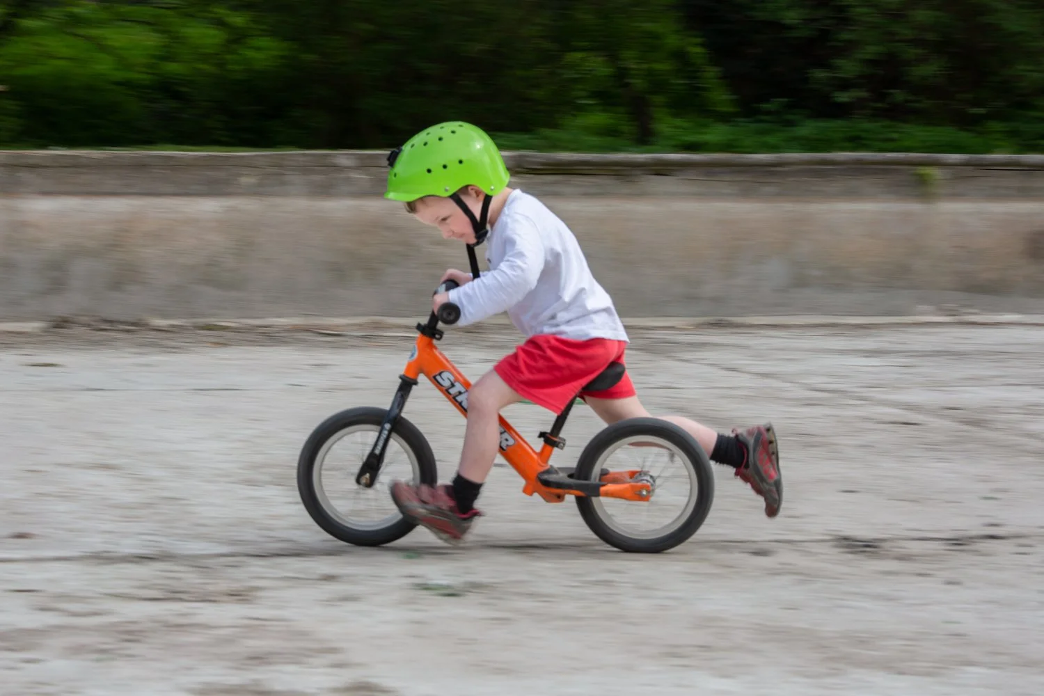 A young boy riding an orange balance bike with a green helmet on a gravel path outdoors.