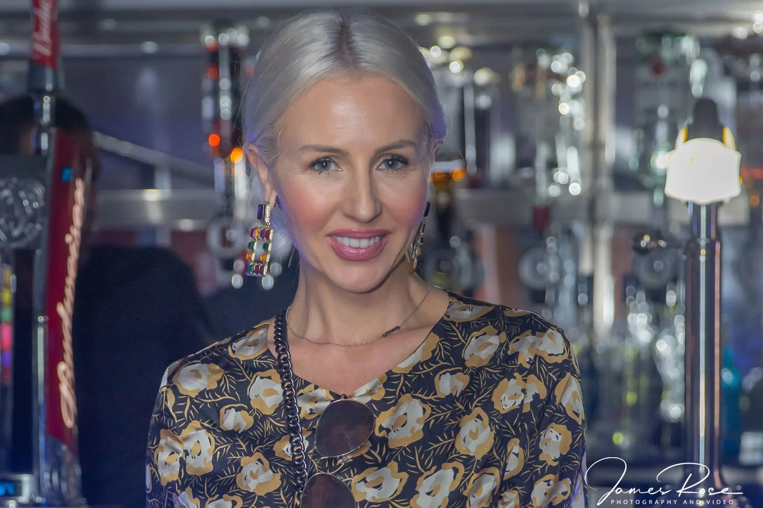 A woman with short blond hair, wearing a black and beige floral dress, colorful earrings, and sunglasses hanging from her shirt, smiling at the camera in an indoor setting with metallic structures and blurred lights in the background.