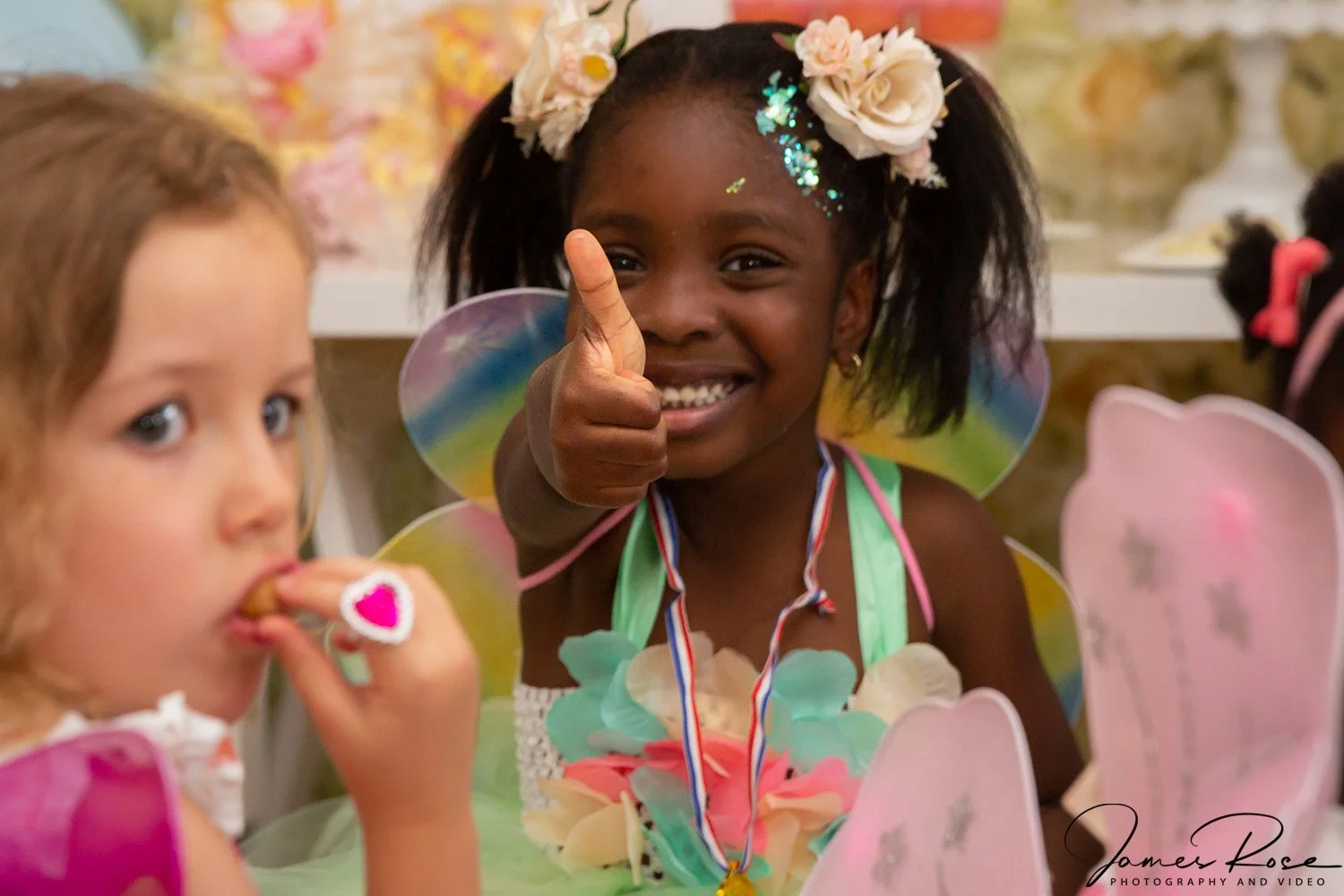 A young girl in a colorful fairy costume giving a thumbs-up at a party, with a bright smile on her face. She is wearing a flower crown, rainbow-colored wings, and a flower-decorated dress. Another girl with curly hair is in the foreground, eating a t