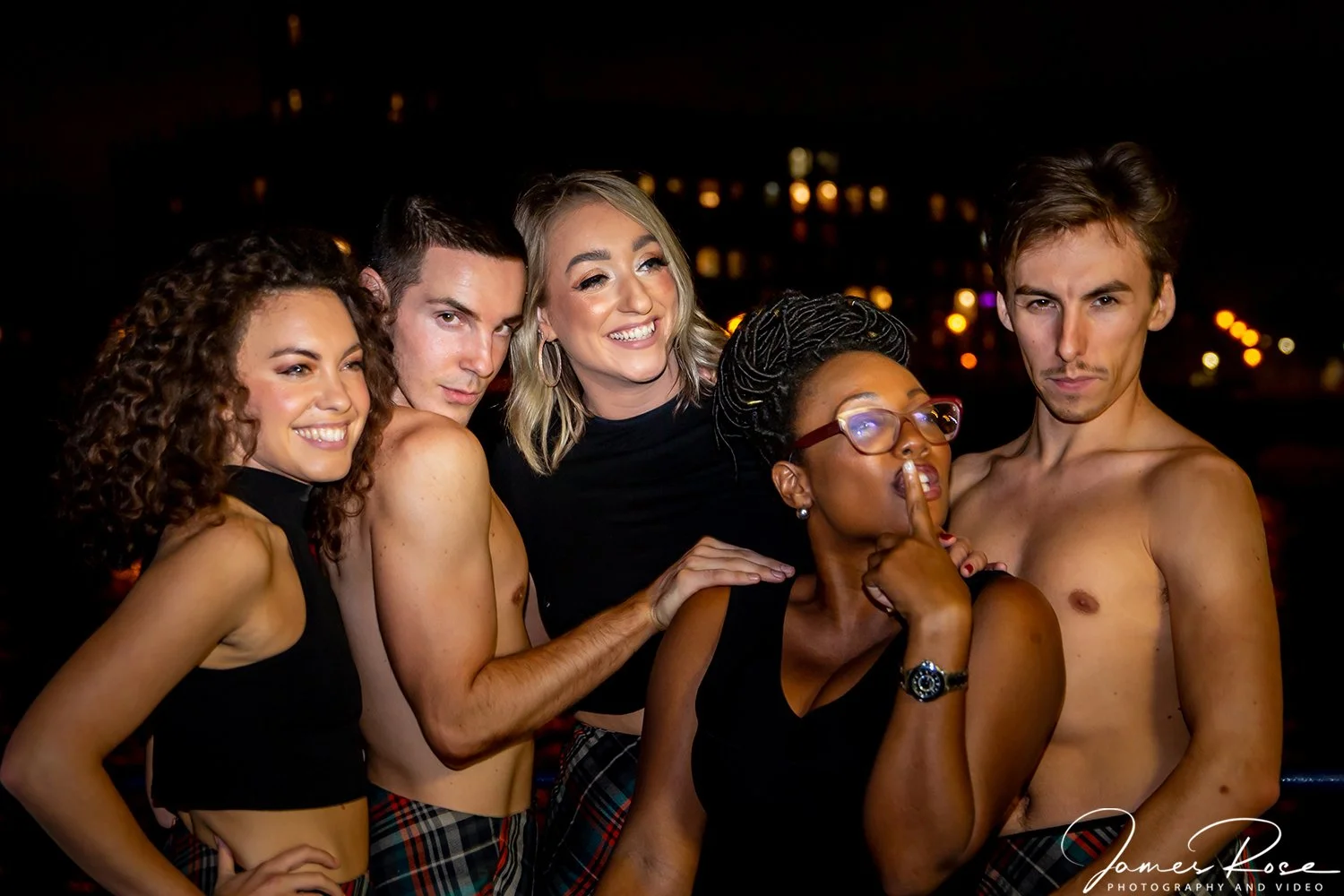 Group of five young adults at night, some shirtless, posing together outdoors with city lights in the background.