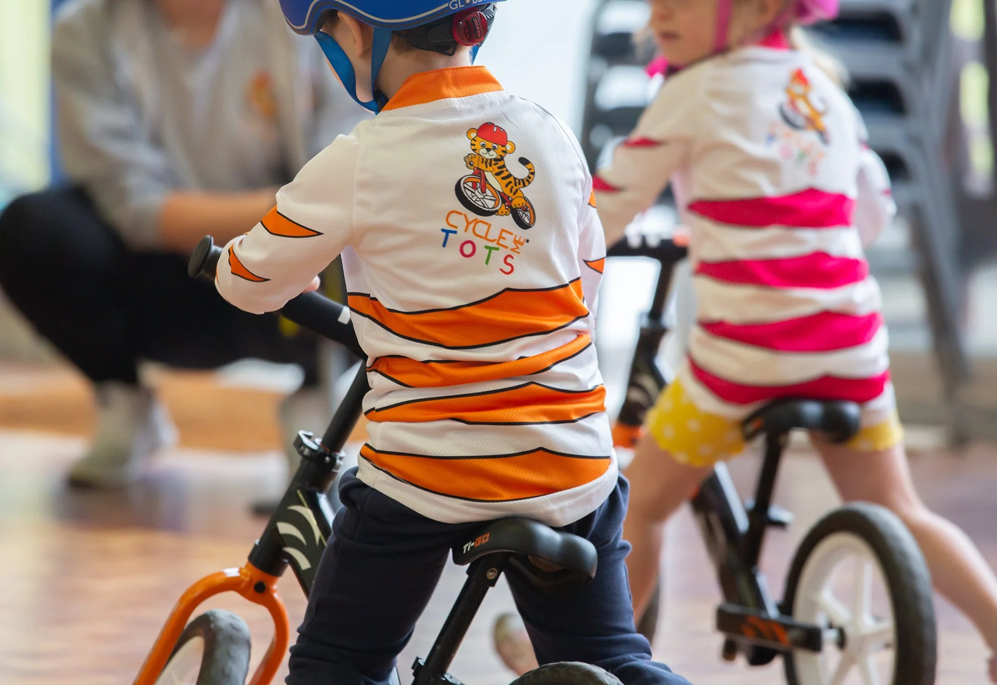 Children wearing colorful striped shirts riding balance bikes indoors, with a person sitting in the background.
