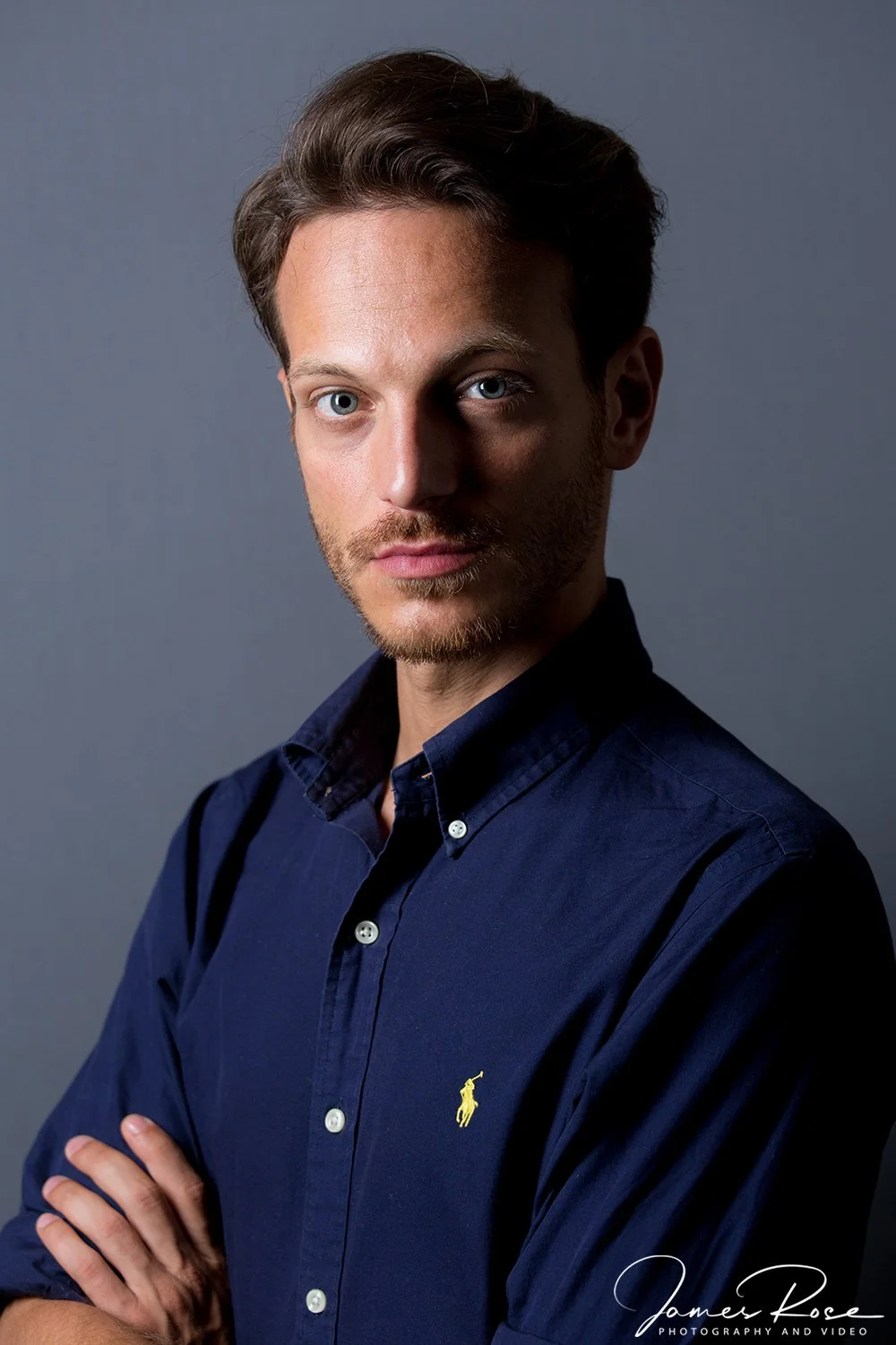 A young man with blue eyes and light brown hair, wearing a dark blue button-up shirt with a yellow Polo Ralph Lauren logo, poses with arms crossed against a plain gray background.