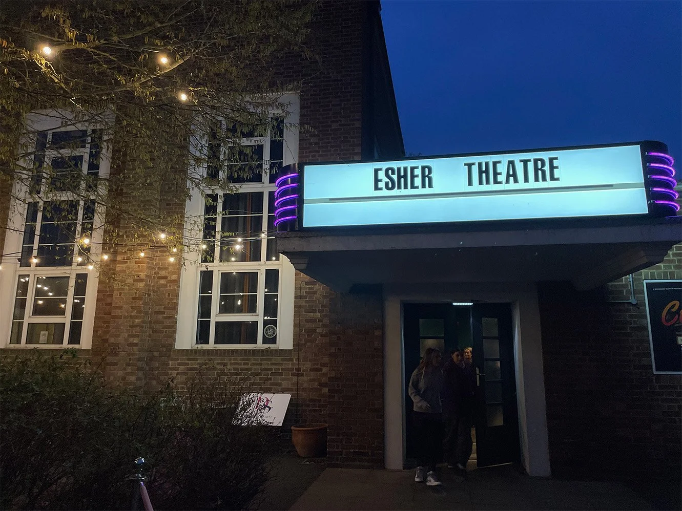 Marquee sign displaying 'Esher Theatre' outside a brick building, with string lights, a woman and a man near the entrance, and illuminated windows at night.