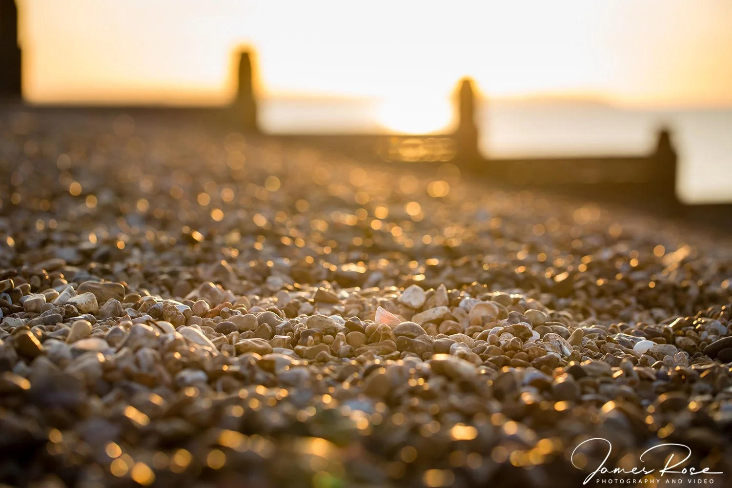 Close-up of pebbles on a beach at sunset with a wooden pier in the background, blurred, during golden hour.