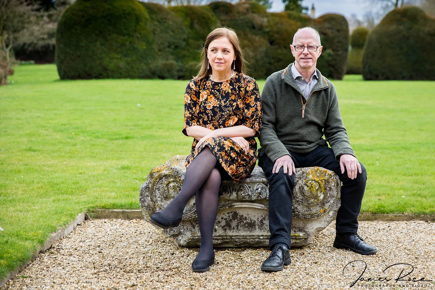 A woman and a man sitting on an ornate stone bench in a garden with green grass and large trimmed bushes in the background.
