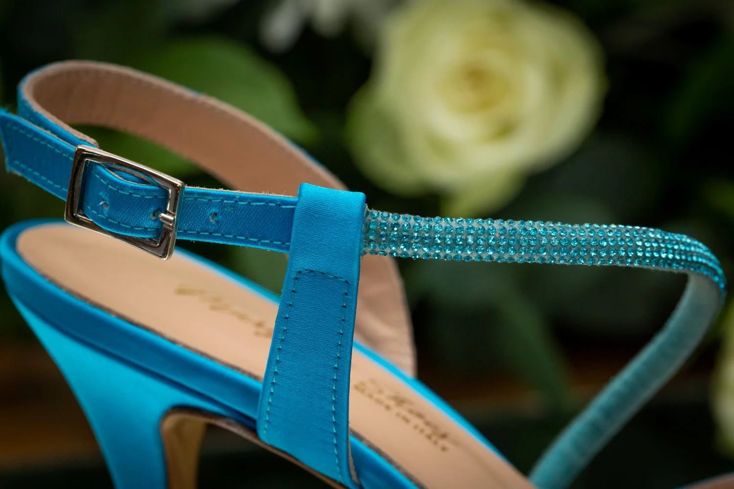 Close-up of a blue high-heeled sandal with rhinestone-embellished ankle strap, set against a blurred green flower background.