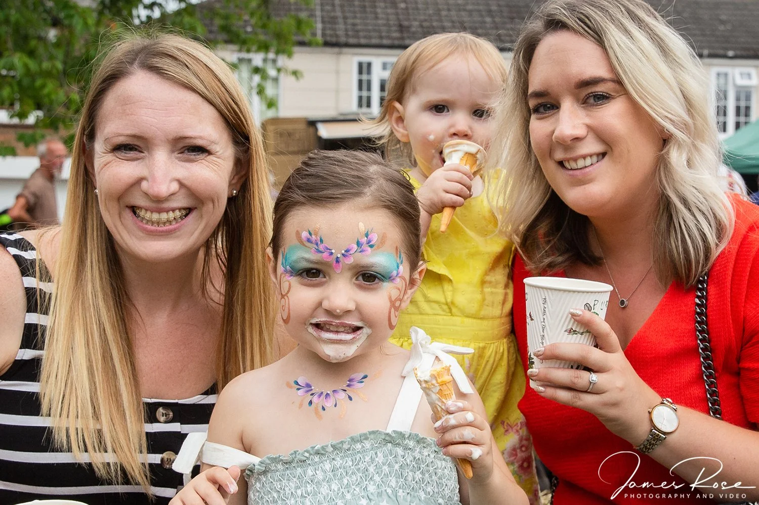 Two women and two children enjoying ice cream at an outdoor gathering, with one girl having face paint and frosting on her face.