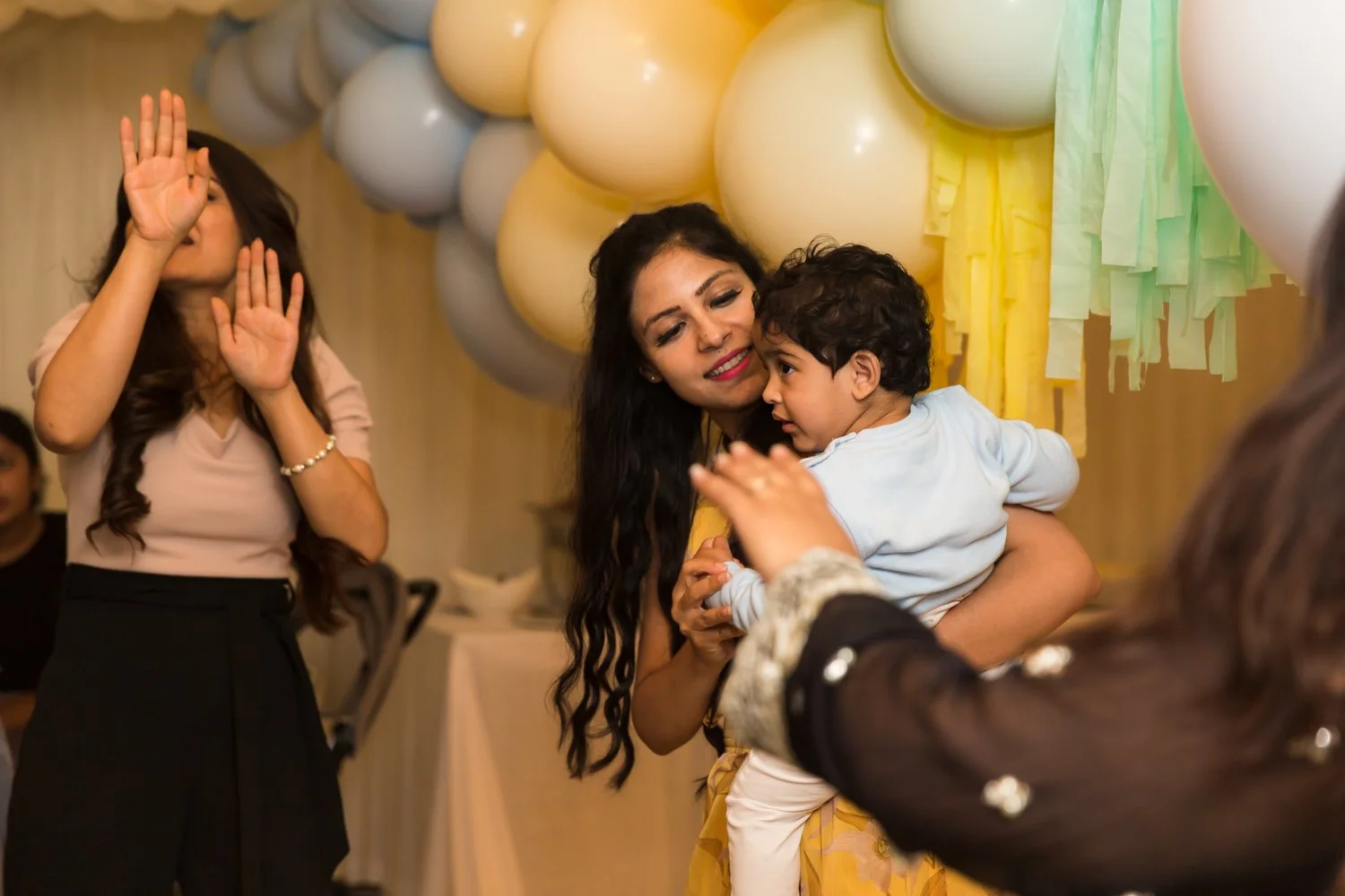 A woman holding a young child in a celebration with balloons and decorative paper and balloons in the background, while another woman appears to be dancing or clapping.