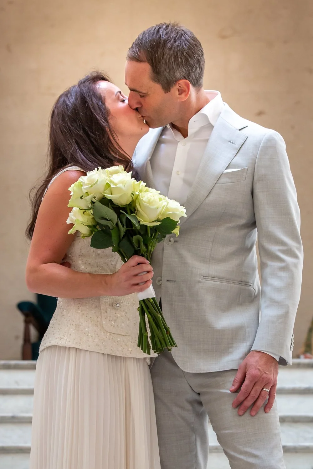 A bride and groom sharing a kiss at their wedding, with the bride holding a bouquet of white roses, both dressed in wedding attire.